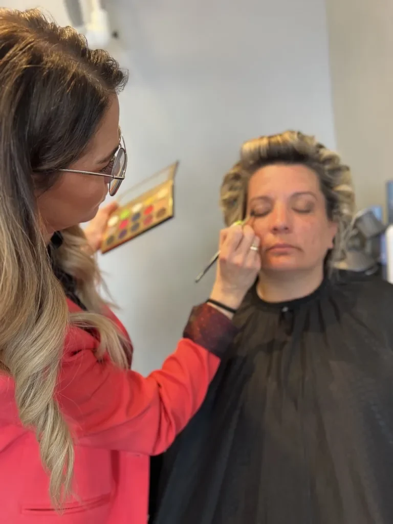 Brudespecialist står med makeuppalletten der fuldender brundes smukke øjenskygge. bruden sidder med lukkede øjne og nyder stylingen i tryghed og ro