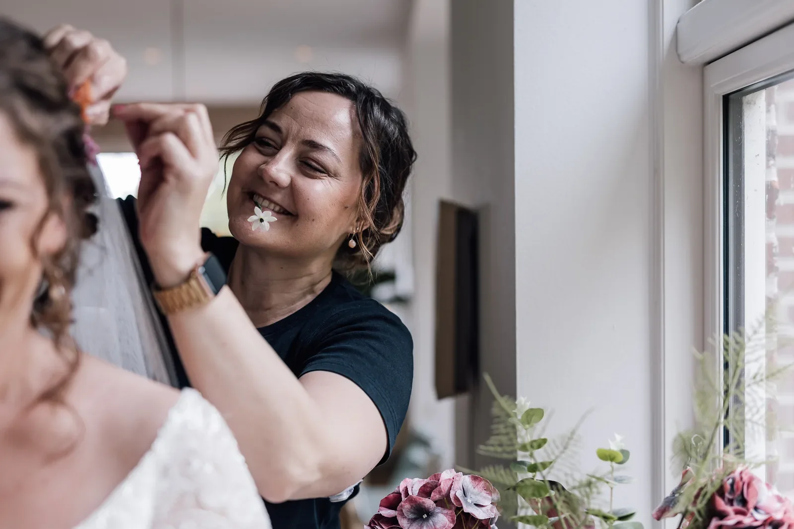 Brudestylist og stifter af Riistyle, Rikke Riis står med en blomst i munden imens hun sætter sløret i håret på bruden i forgrunden