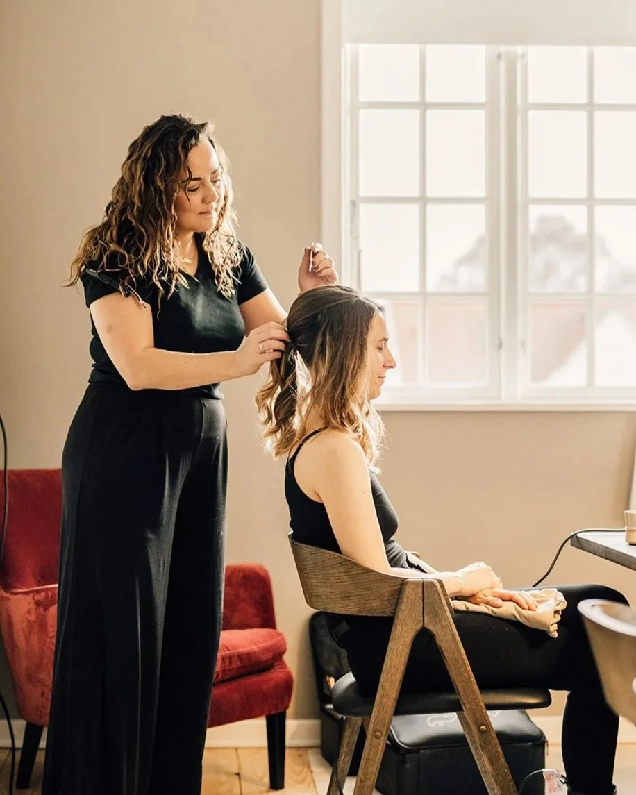 Brudestylist Rikke står bag en brud som sidder med lukkede øjne og nyder roen. Stylingen foregår foran stort lyst vidue og med en rød læne stol i baggrunden. Stemningen er rolig og hjemnin