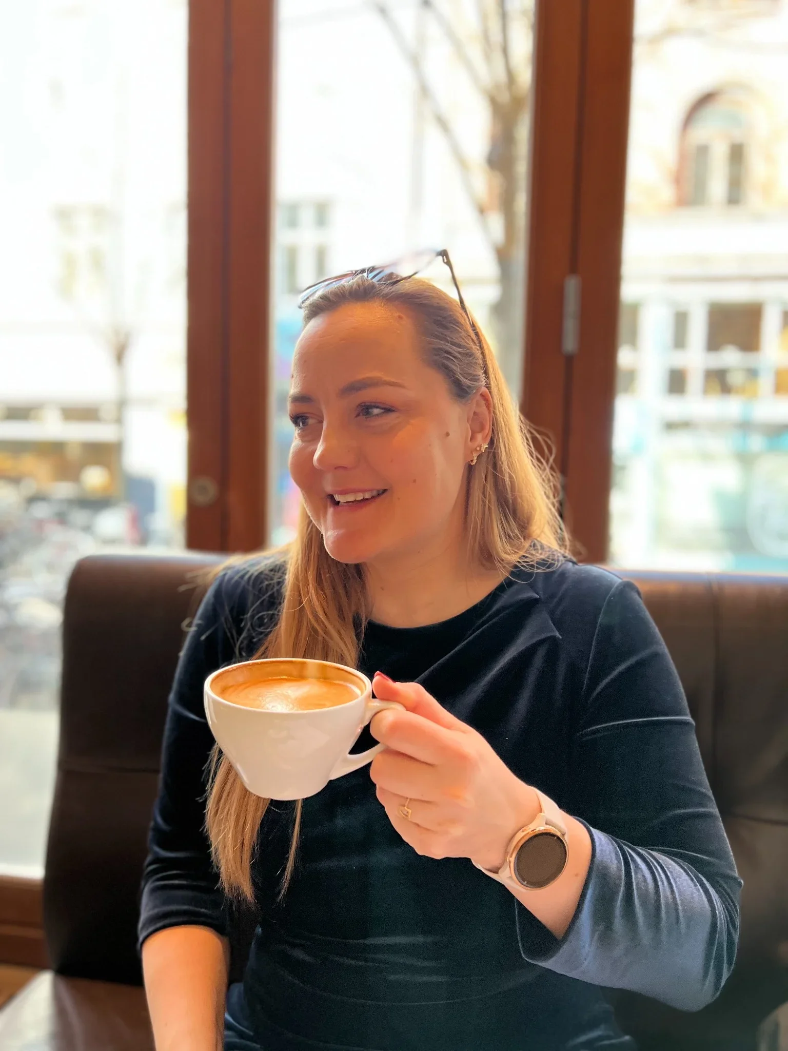 Rikke fra RiiStyle sidder med en kop kaffe i hånden og smiler i et roligt café-miljø, et varmt og personligt øjeblik der viser hendes nærvær og naturlige energi.