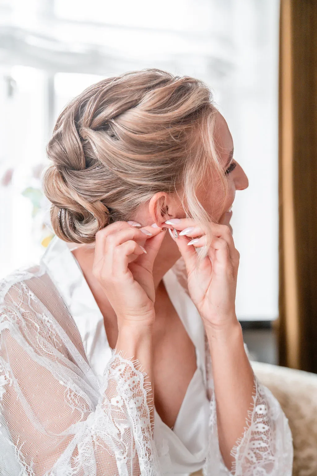Brud gør sig klar og tager øreringe på med elegant flettet håropsætning – romantisk og naturligt brudehår i skandinavisk stil.