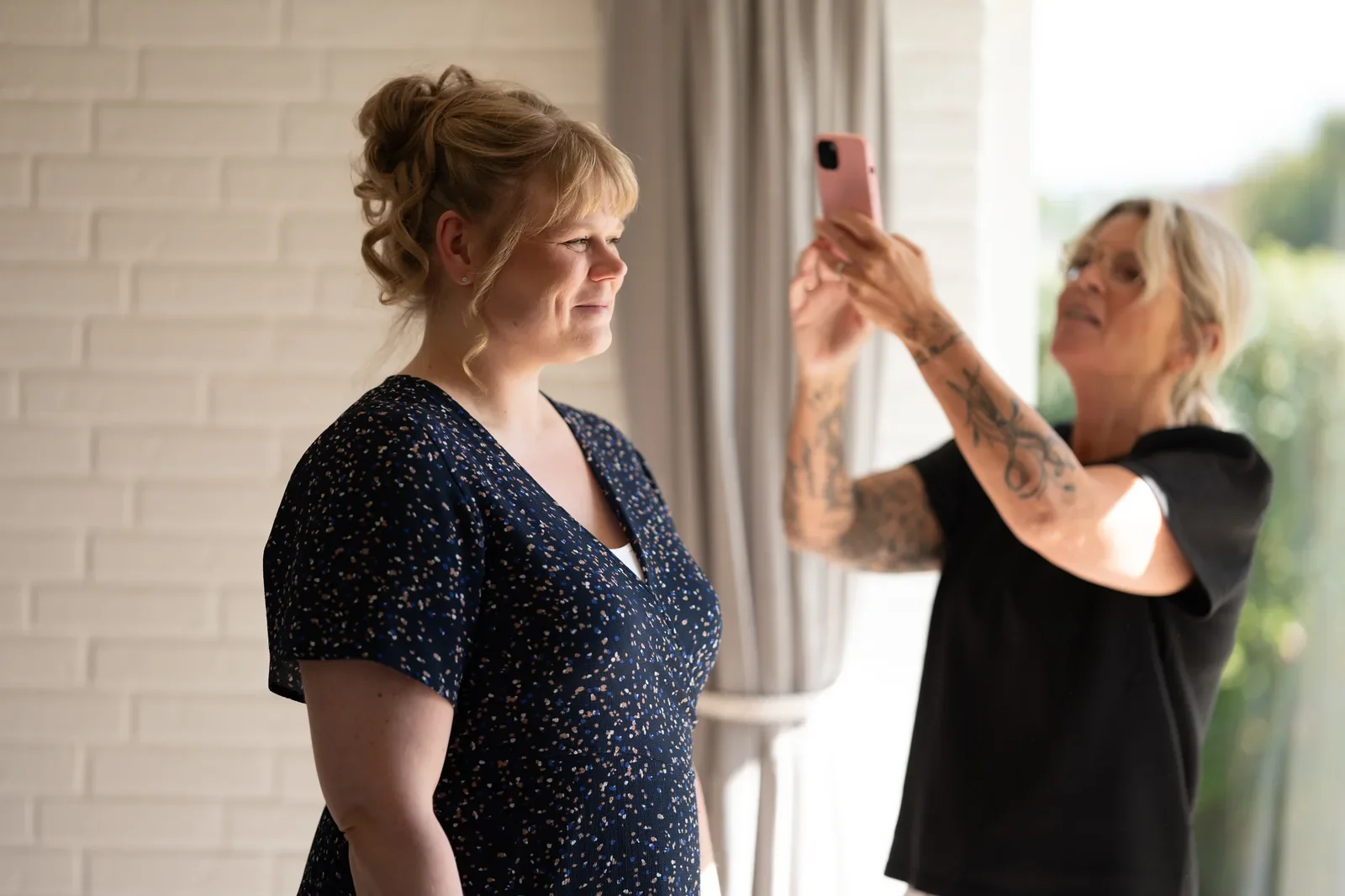 i forgrunden en glad mor til bruden som har fået sat flot hår og lagt makeup. bagved står brudestylisten og tager billeder af håret med sin telefon