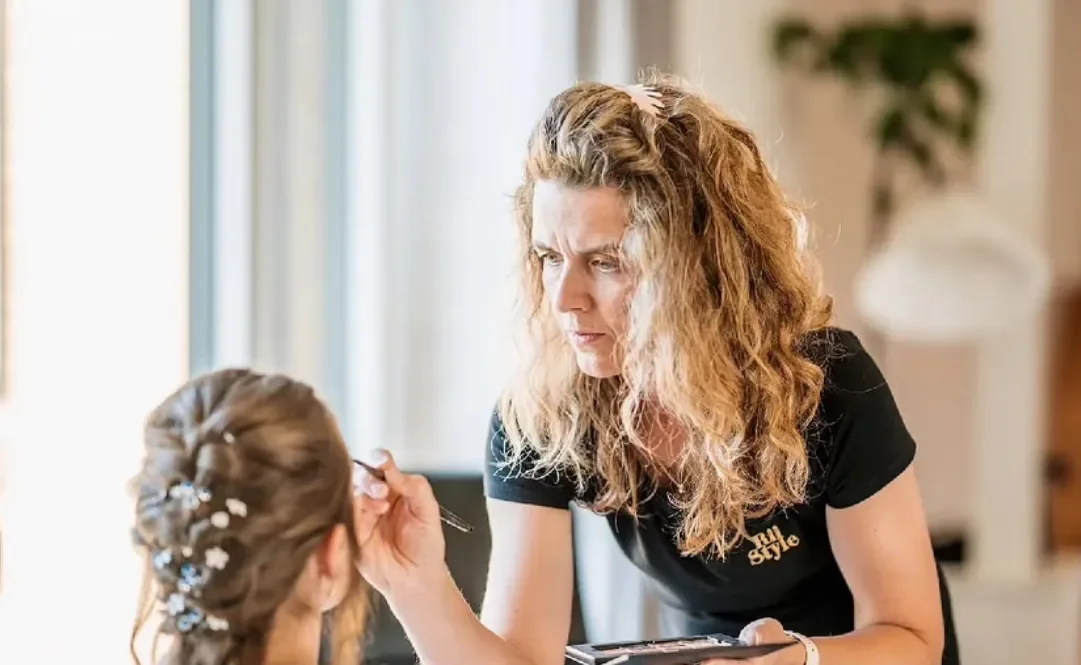 Frisør og brudespecialist står foroverbøjet foran bruden i stolen hun lægger makeup og ser koncentreret ud. Brudens hår er flettet og med perler i håret