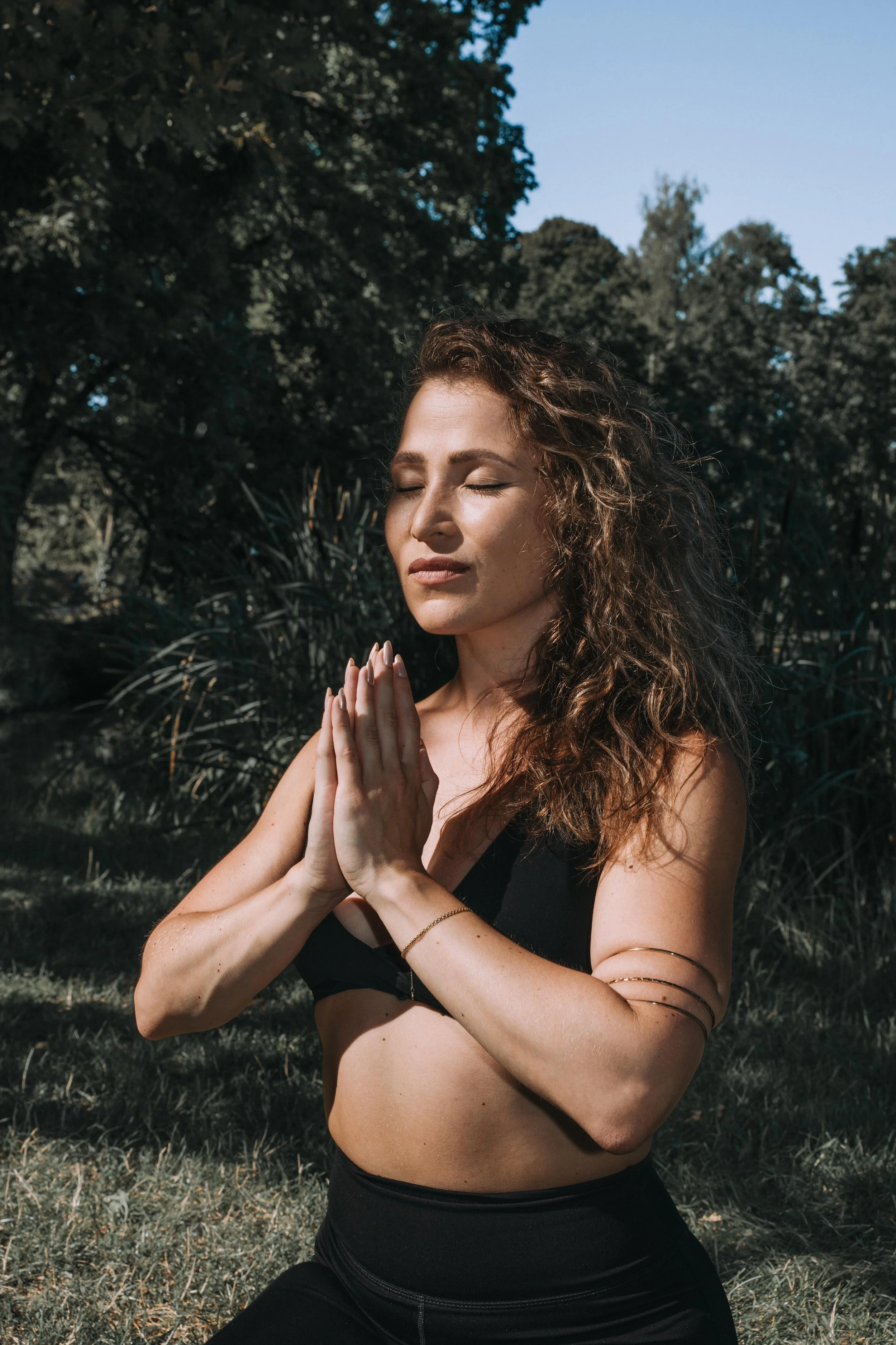Junge Frau praktiziert Yoga im Freien, meditiert mit geschlossenen Augen, in einer grünen, bewaldeten Umgebung unter blauem Himmel.