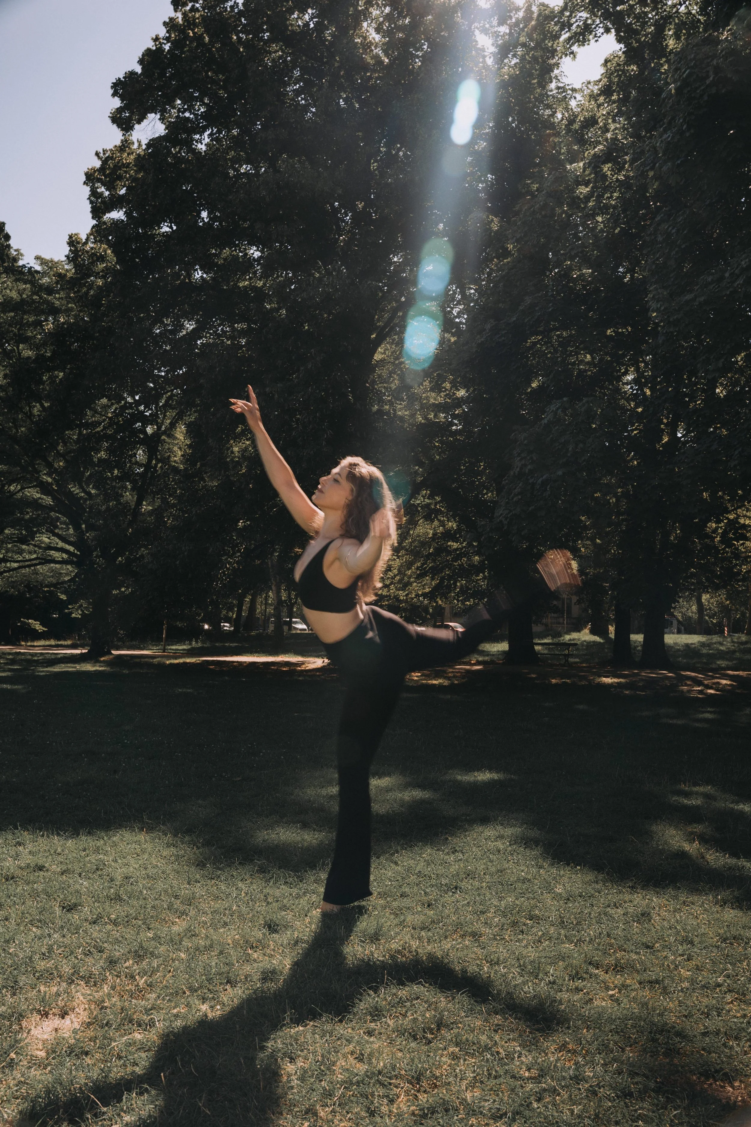 Eine junge Frau steht auf einer Wiese, macht Yoga bei Sonnenlicht, umgeben von Bäumen.