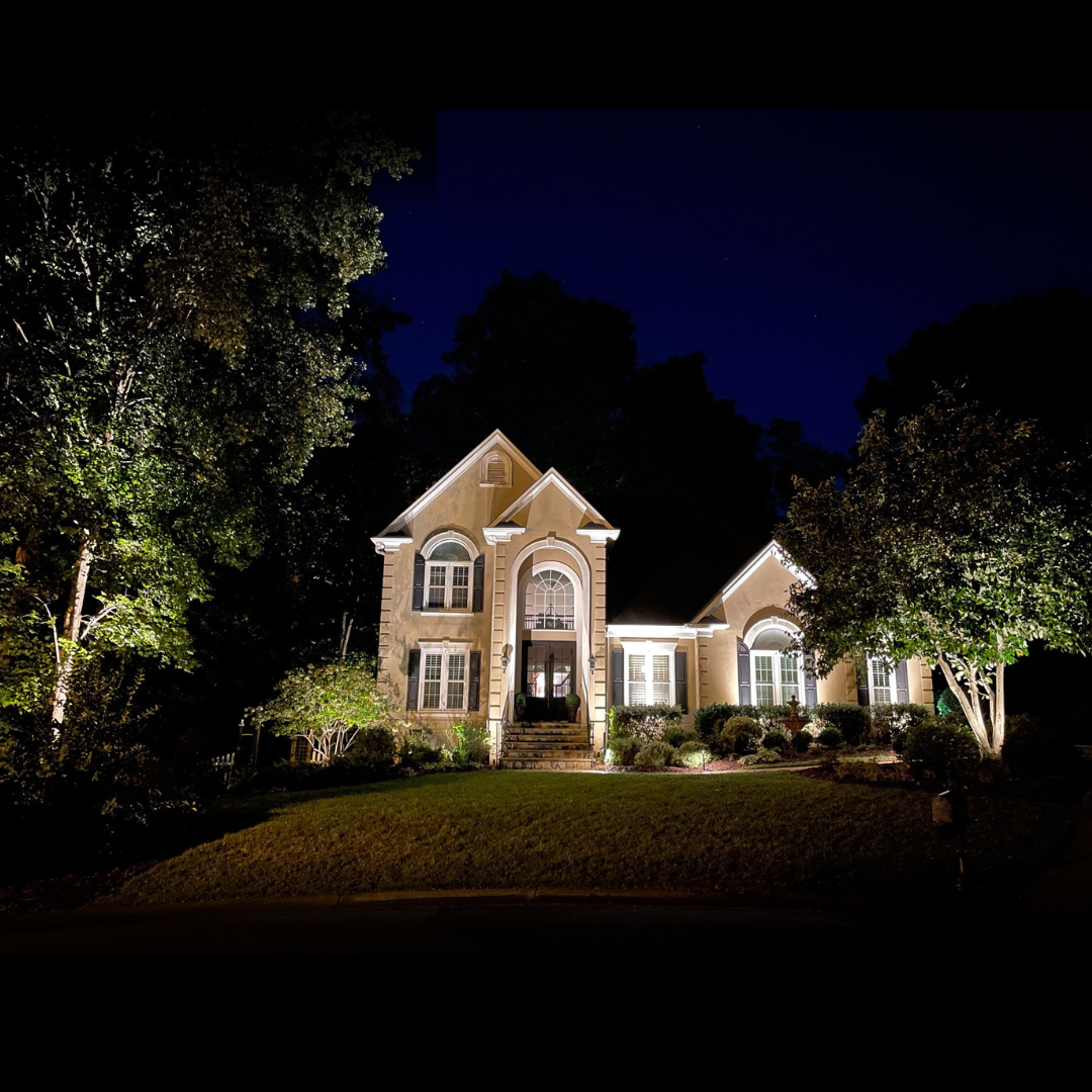  Tan stucco home with dental moulding illuminated with a combination of landscape lighting and architectural up lights. 