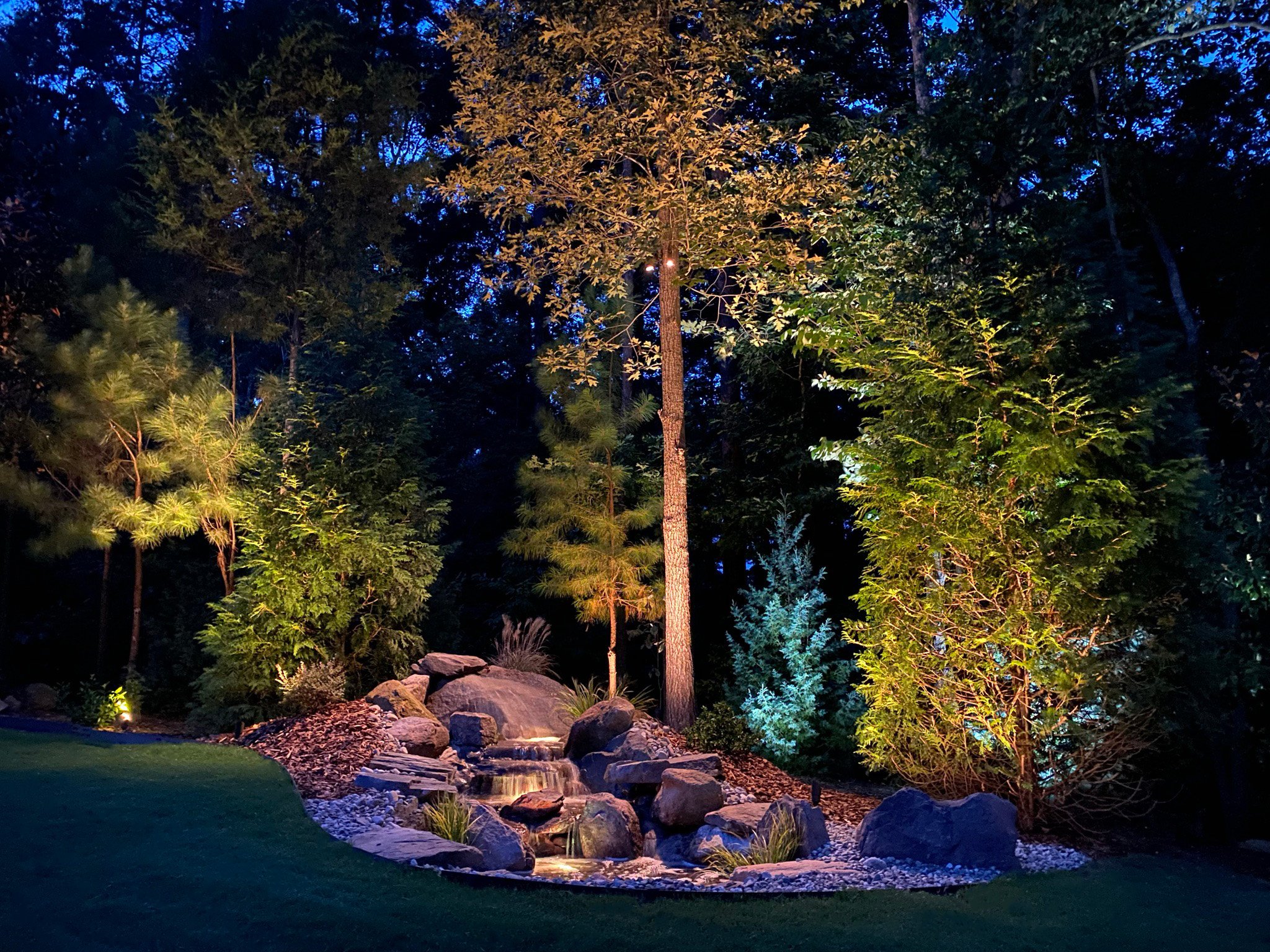  Water feature with a small waterfall surrounded by landscaping rocks is illuminated with underwater lights and down lighting. Surrounding trees are lit with tree accent lighting. 