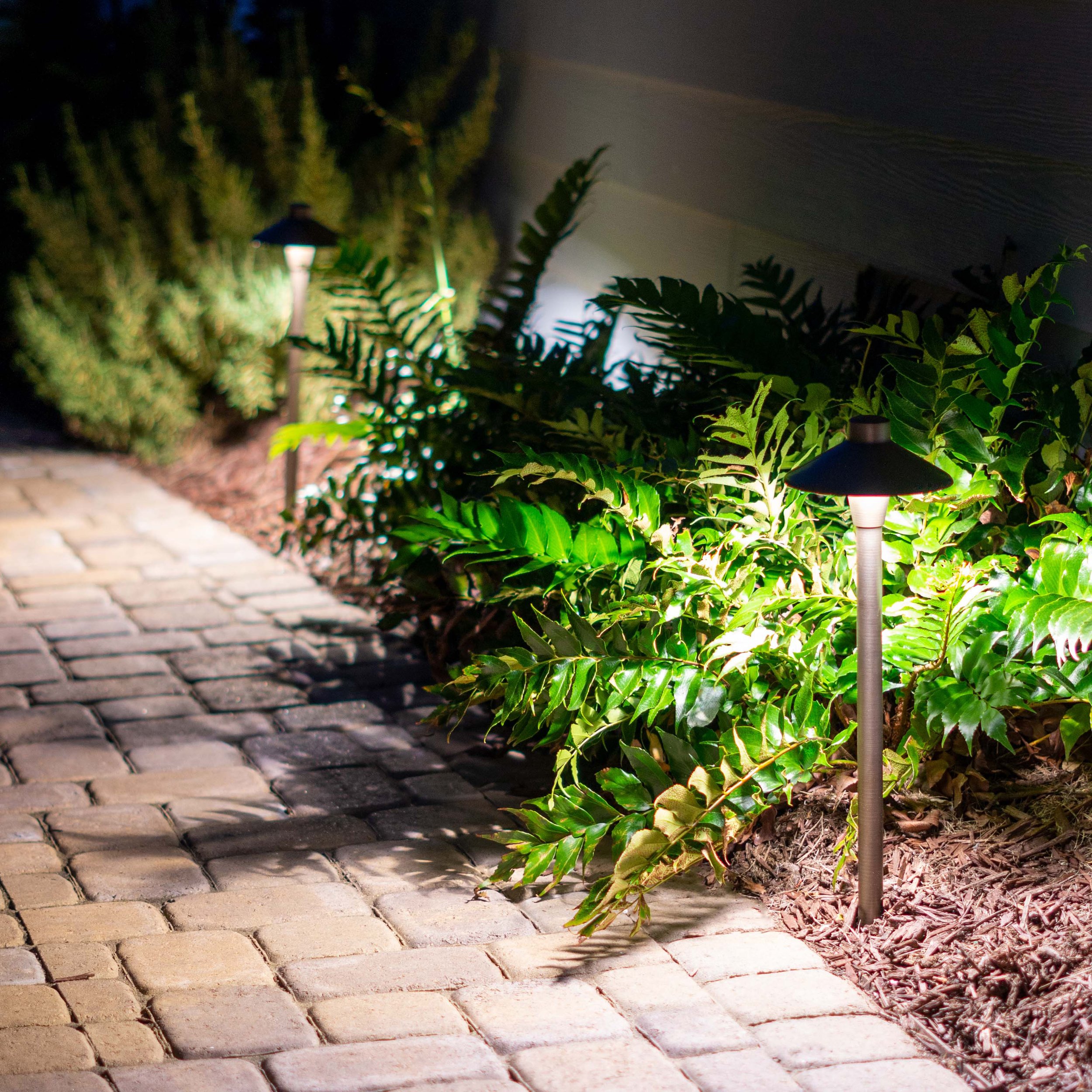  Two traditional path lights illuminate a stamped concrete path alongside a landscaping bed with ferns surrounded by mulch. 