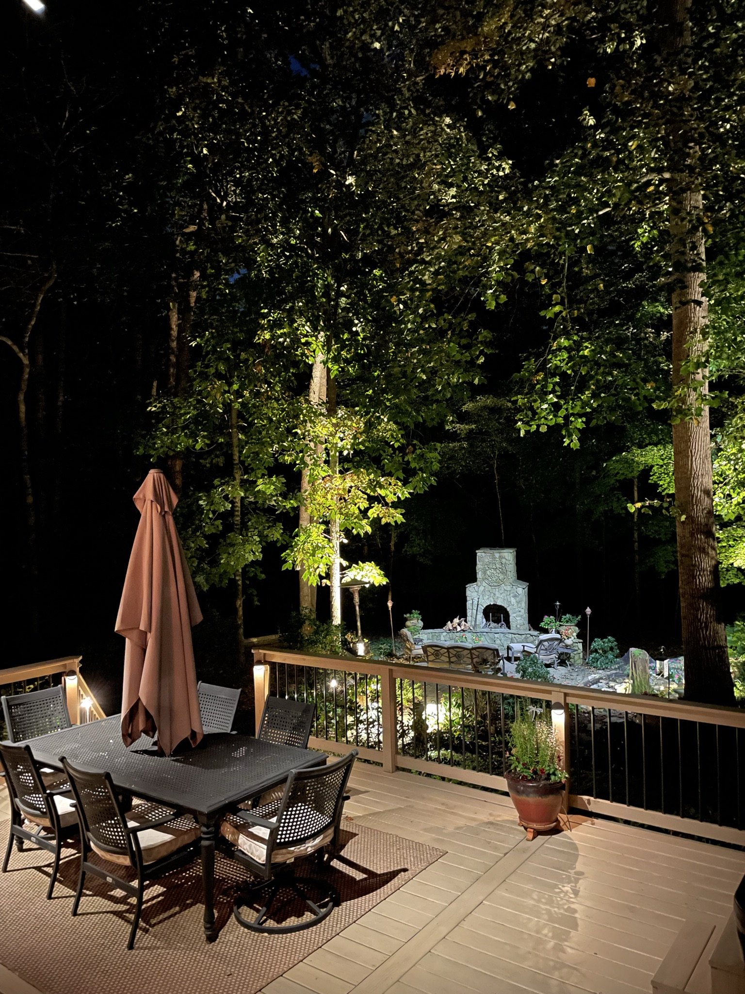  Overhead deck lighting illuminates an outdoor eating area. In the background you can see additional landscape lighting on the trees and outdoor fireplace. 