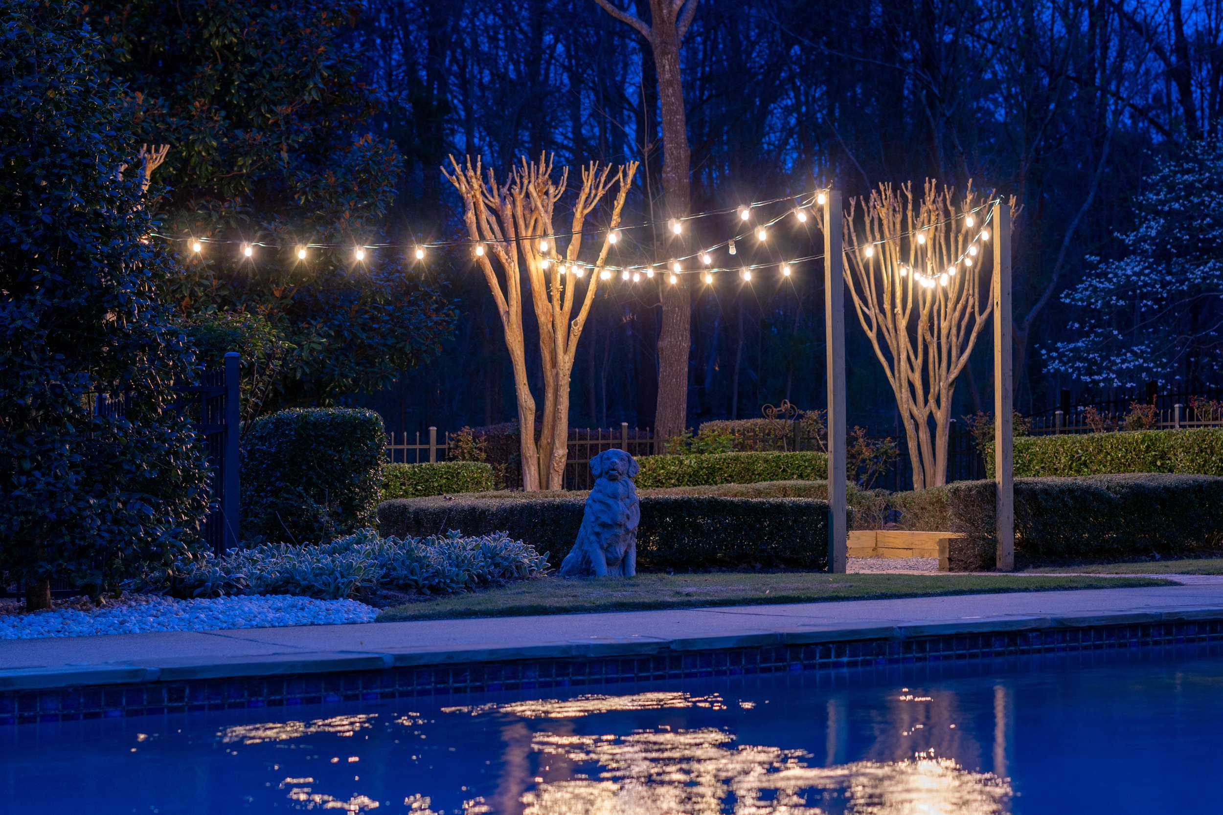  Bistro lights strung between posts and crepe myrtle trees create ambient light for a poolside firepit area. 