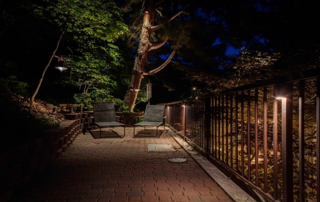  Downlights illuminate a paver patio with a sitting area. Fence lights are also mounted on the fence to the right producing more light on the ground. 