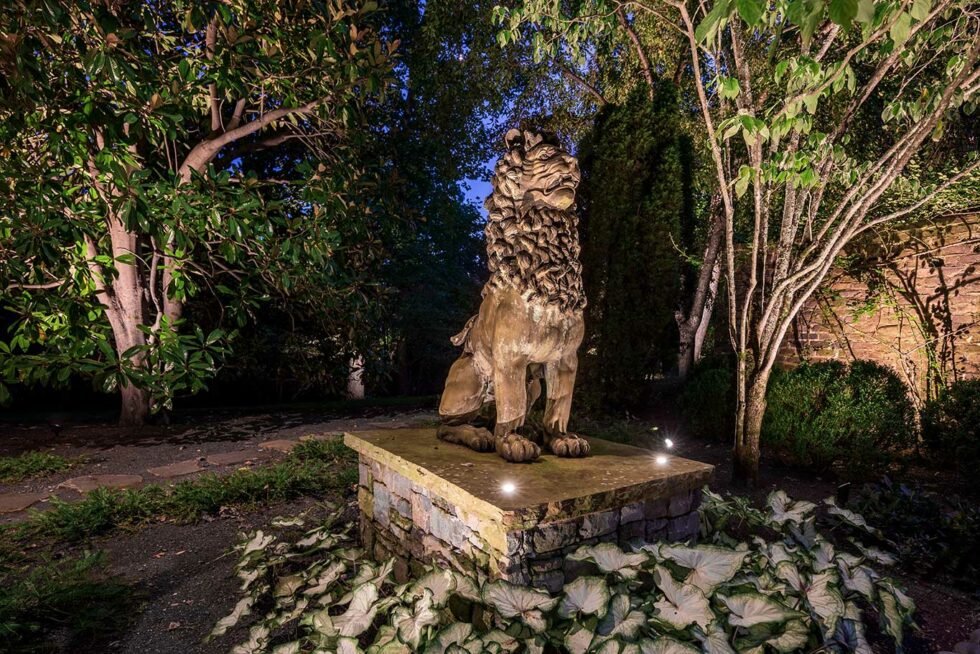  Lion statue situated within a garden is lit with accent lights. Landscaping lights also illuminate the surrounding trees and brick structure in the background. 