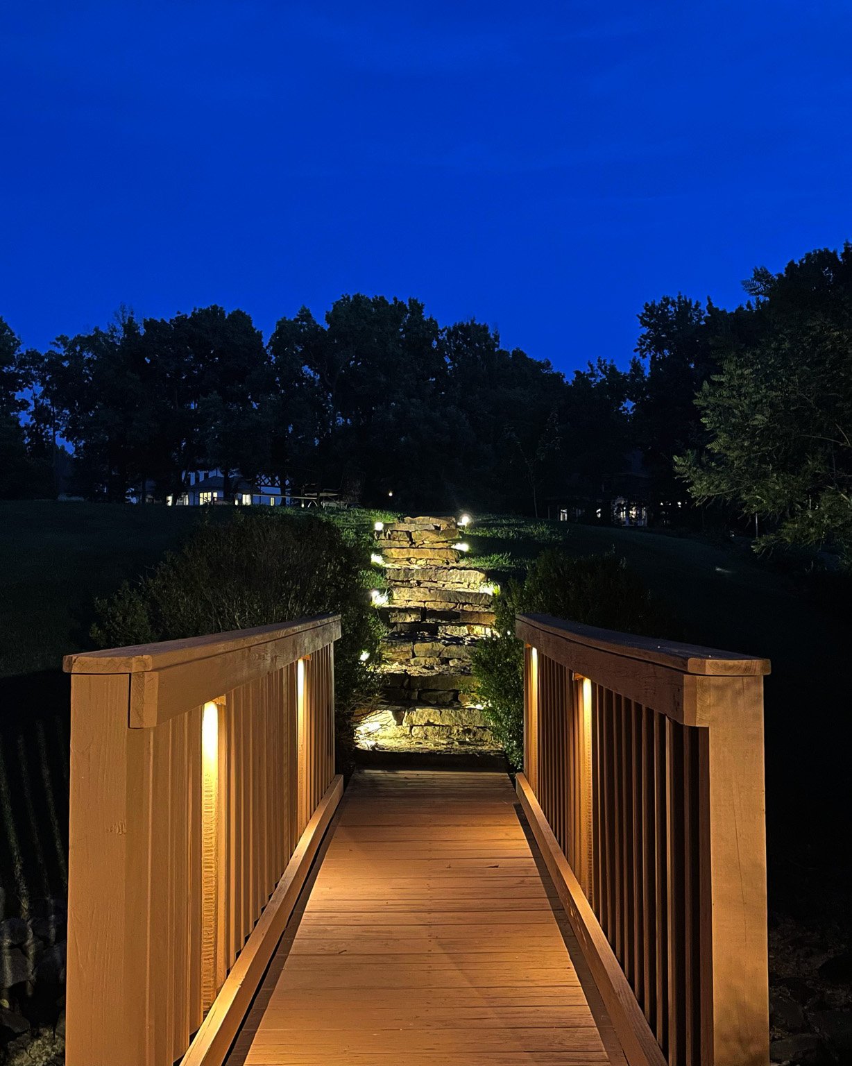  An orange colored wood bridge illuminated by undermount deck lights leads the way to a natural stone path illuminated with low profile path lights. 