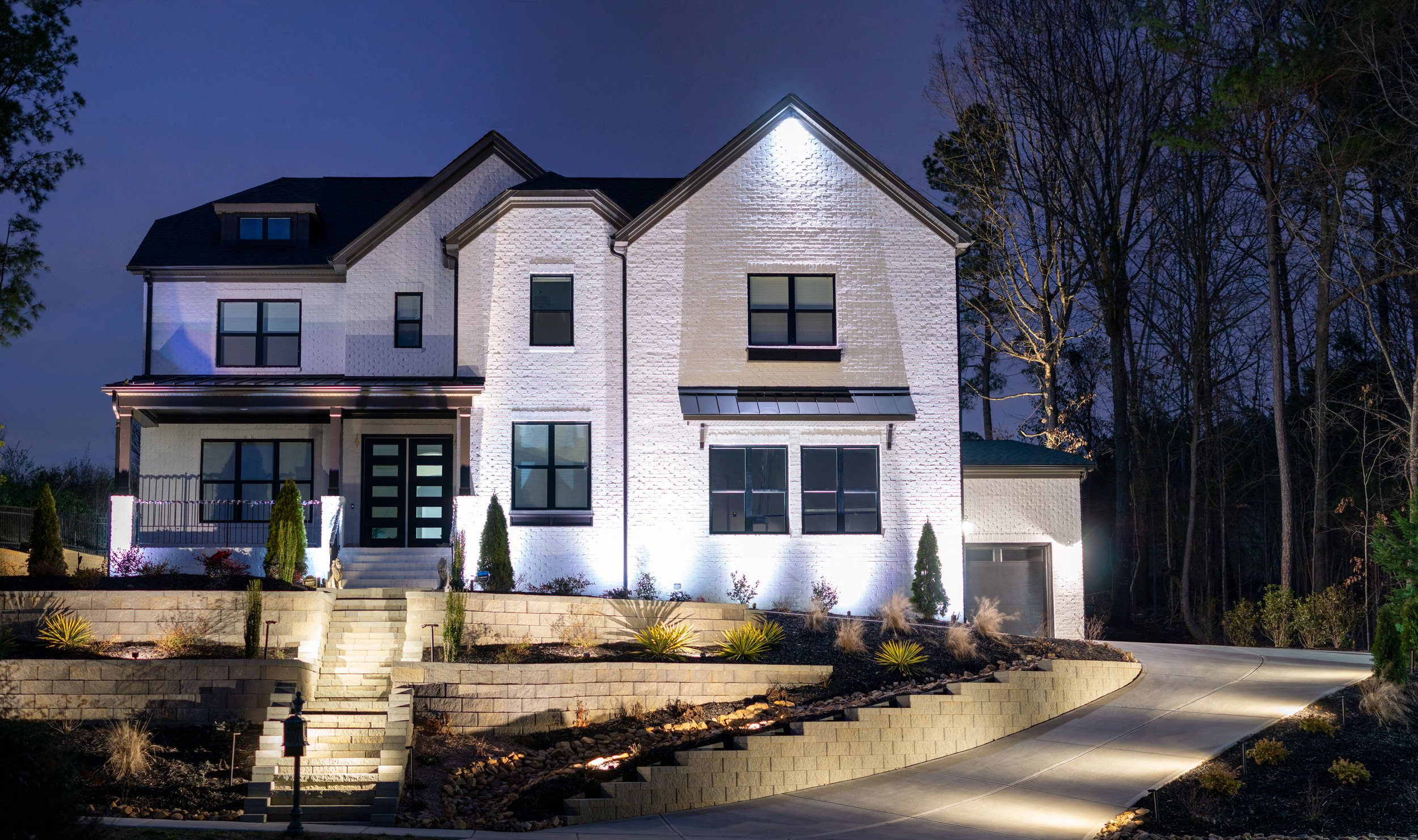  White brick home with dark trim illuminated with architectural up lights and downlights. Front yard is heavily landscaped with tiered retaining walls illuminated with soft landscape lighting. 