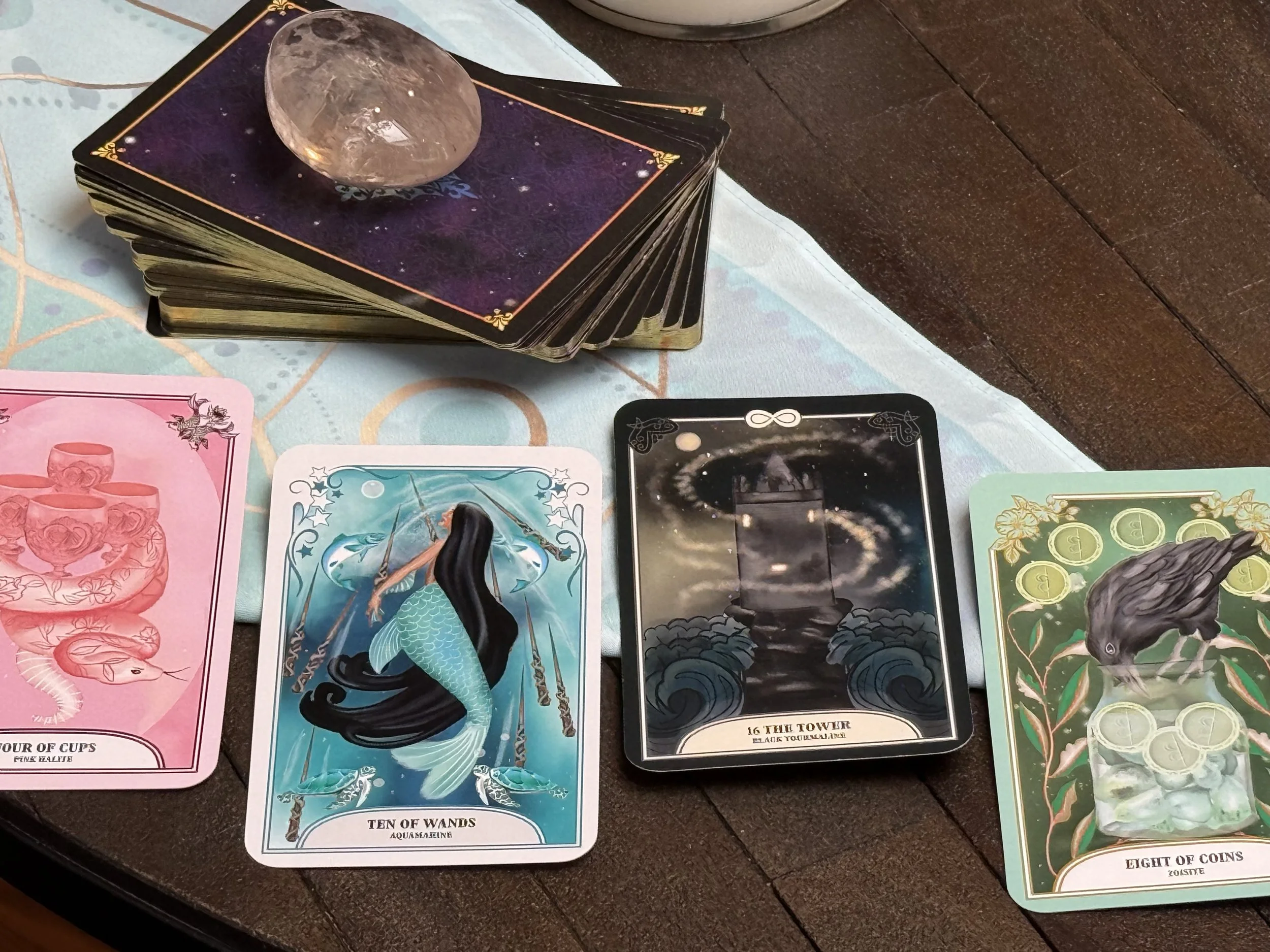 A spread of tarot and oracle cards on a table, with a deck of cards stacked and a crystal on top, surrounded by a cloth with mystical symbols.