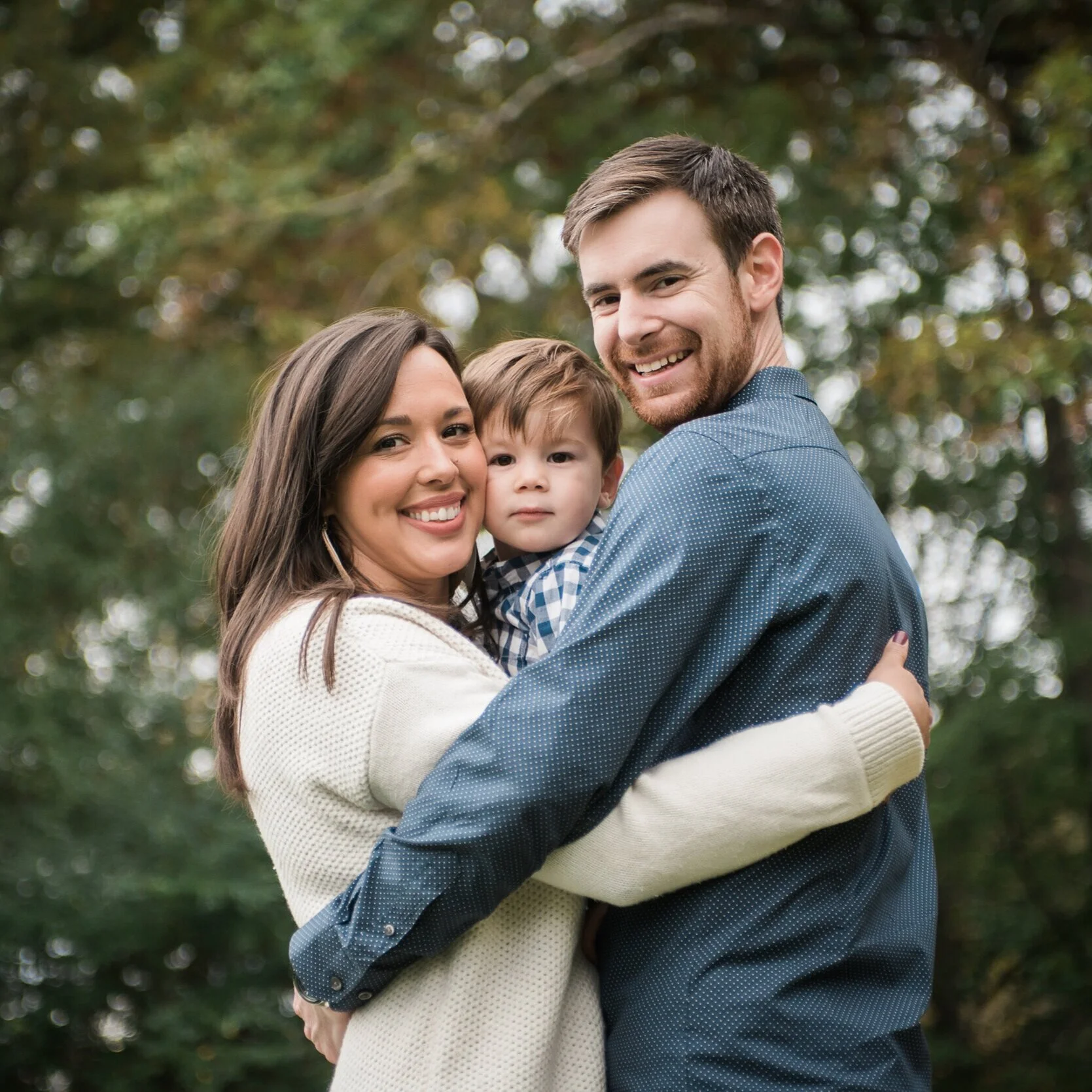 A happy family of three, including a woman, a man, and a small boy, embracing outdoors with trees in the background.