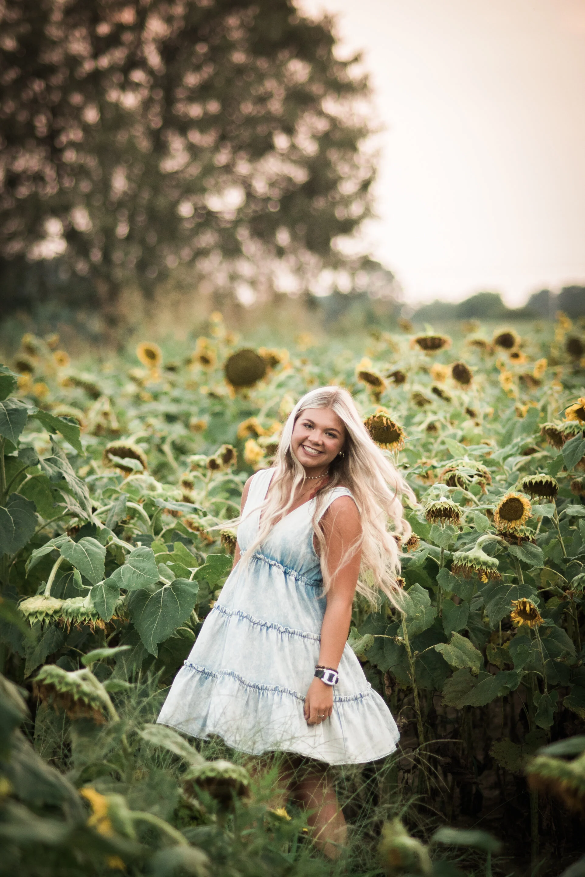 A young woman with long blonde hair smiling and standing in a sunflower field at sunset.