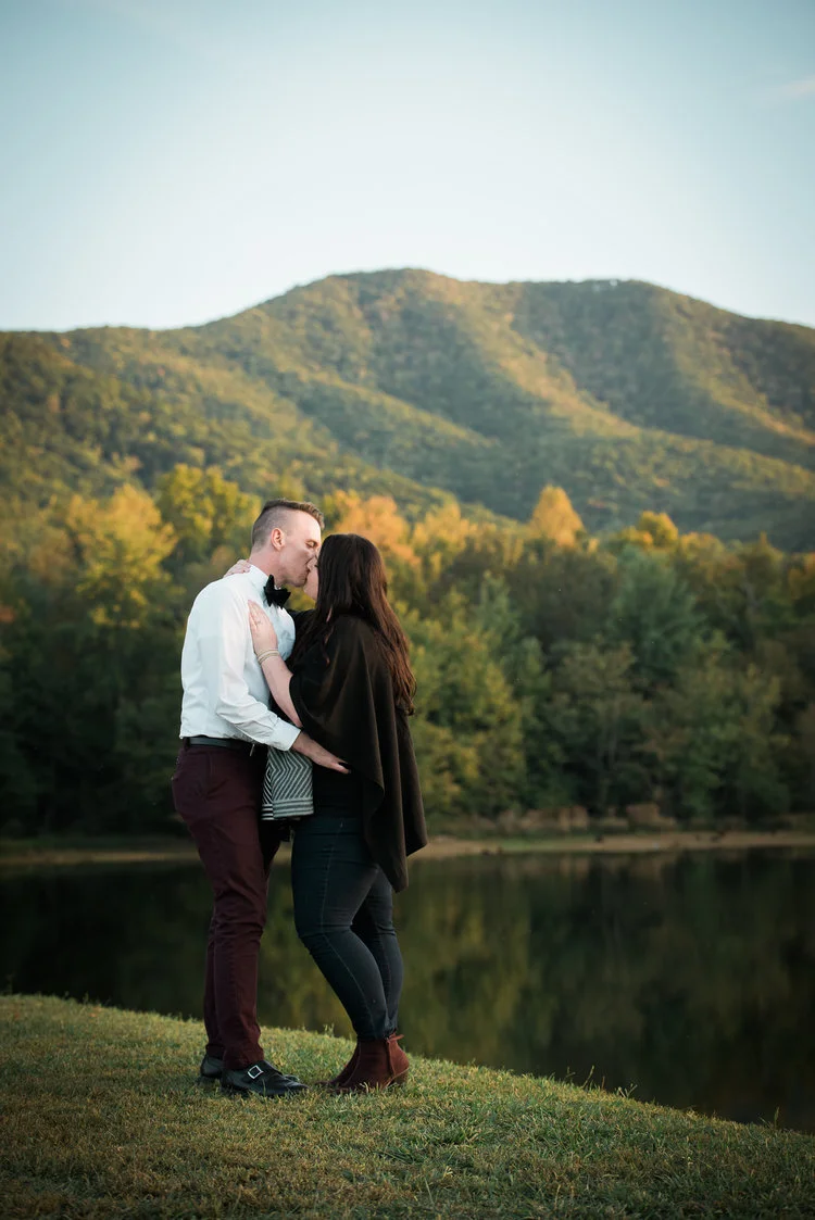 A couple sharing a kiss outdoors near a lake with trees and mountains in the background during the evening.