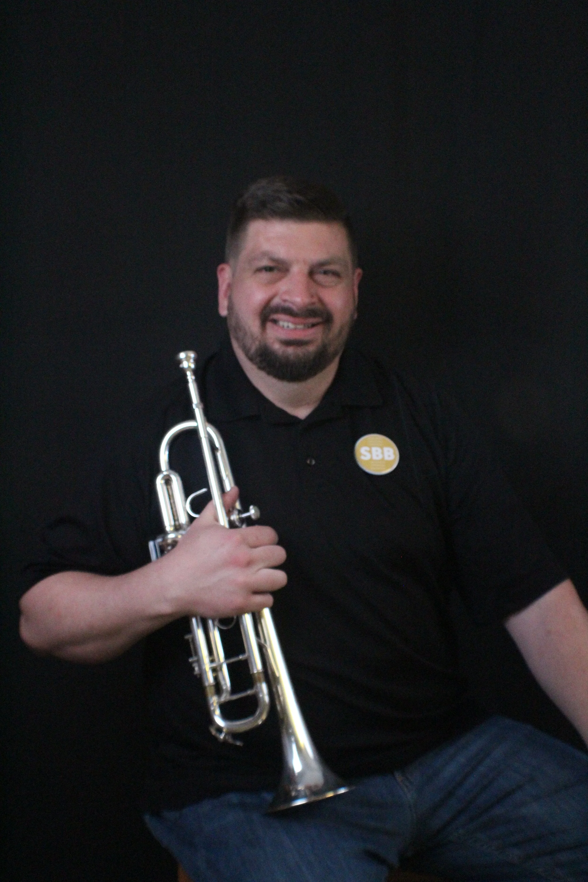 Man with a beard smiling and holding a trumpet, standing against a beige brick wall.
