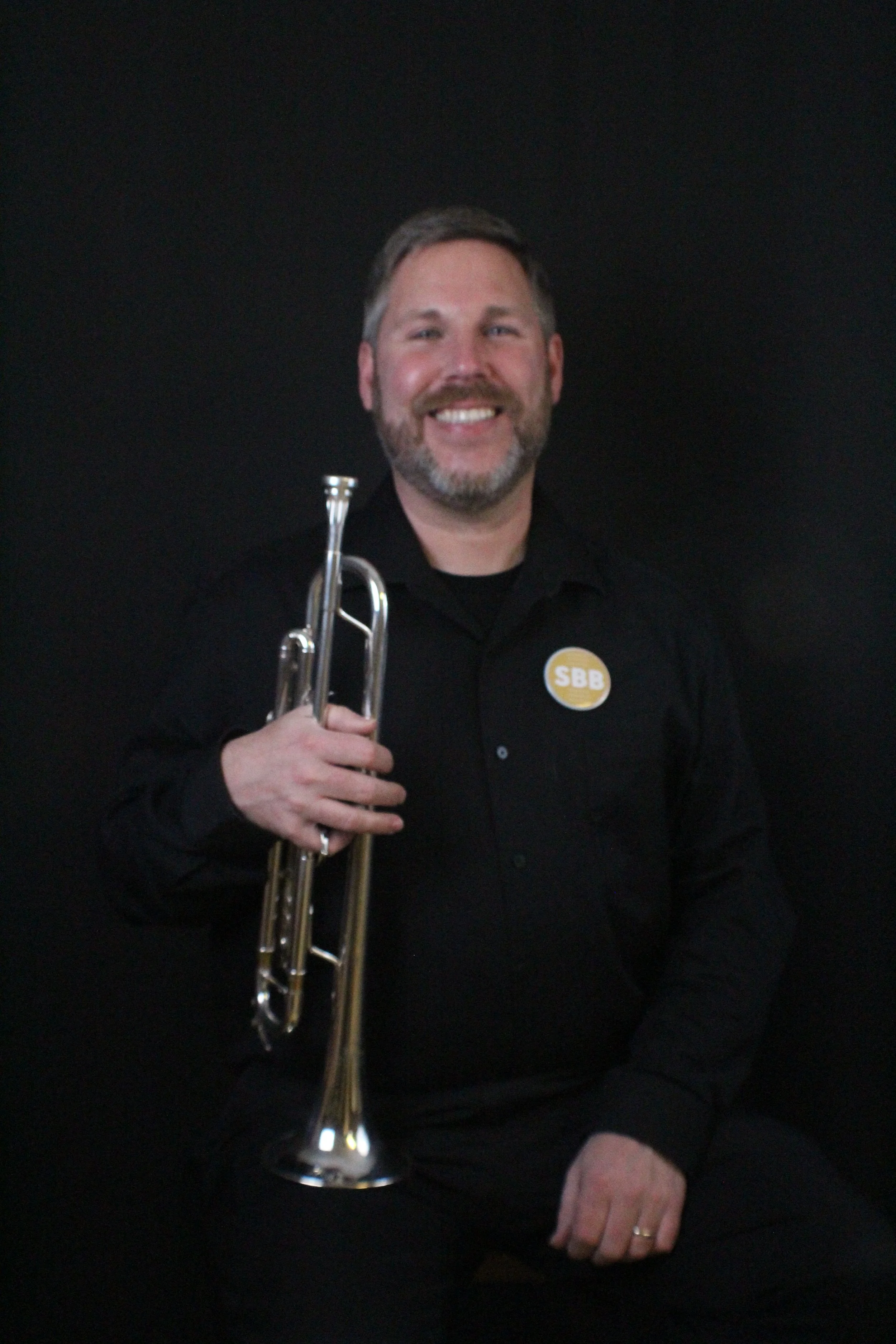 A man with a beard and short hair, wearing a black shirt, smiling and holding a silver trumpet, standing against a light-colored brick wall.