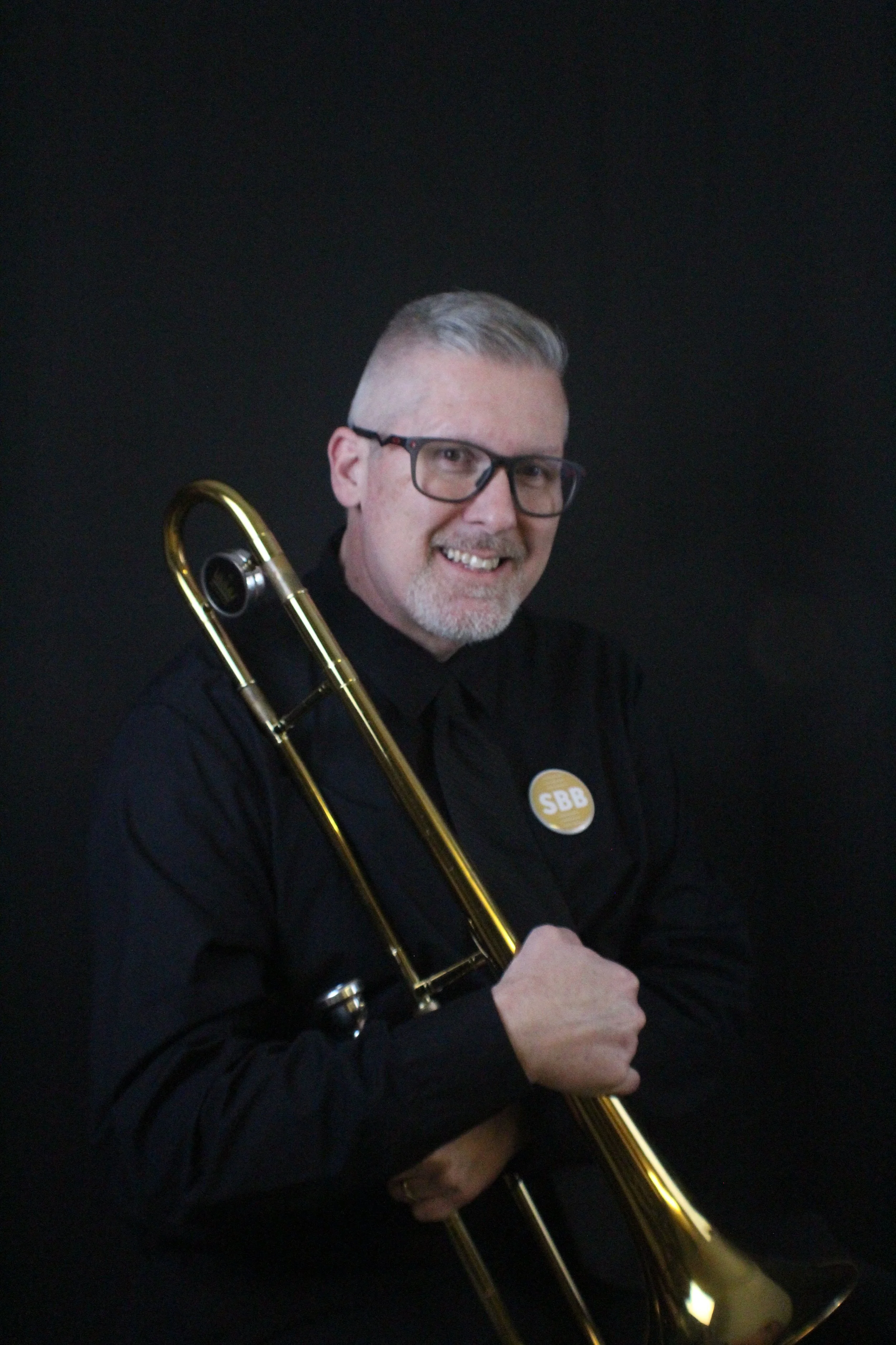 A smiling man with glasses and gray hair, holding a trombone, standing in front of a beige brick wall.