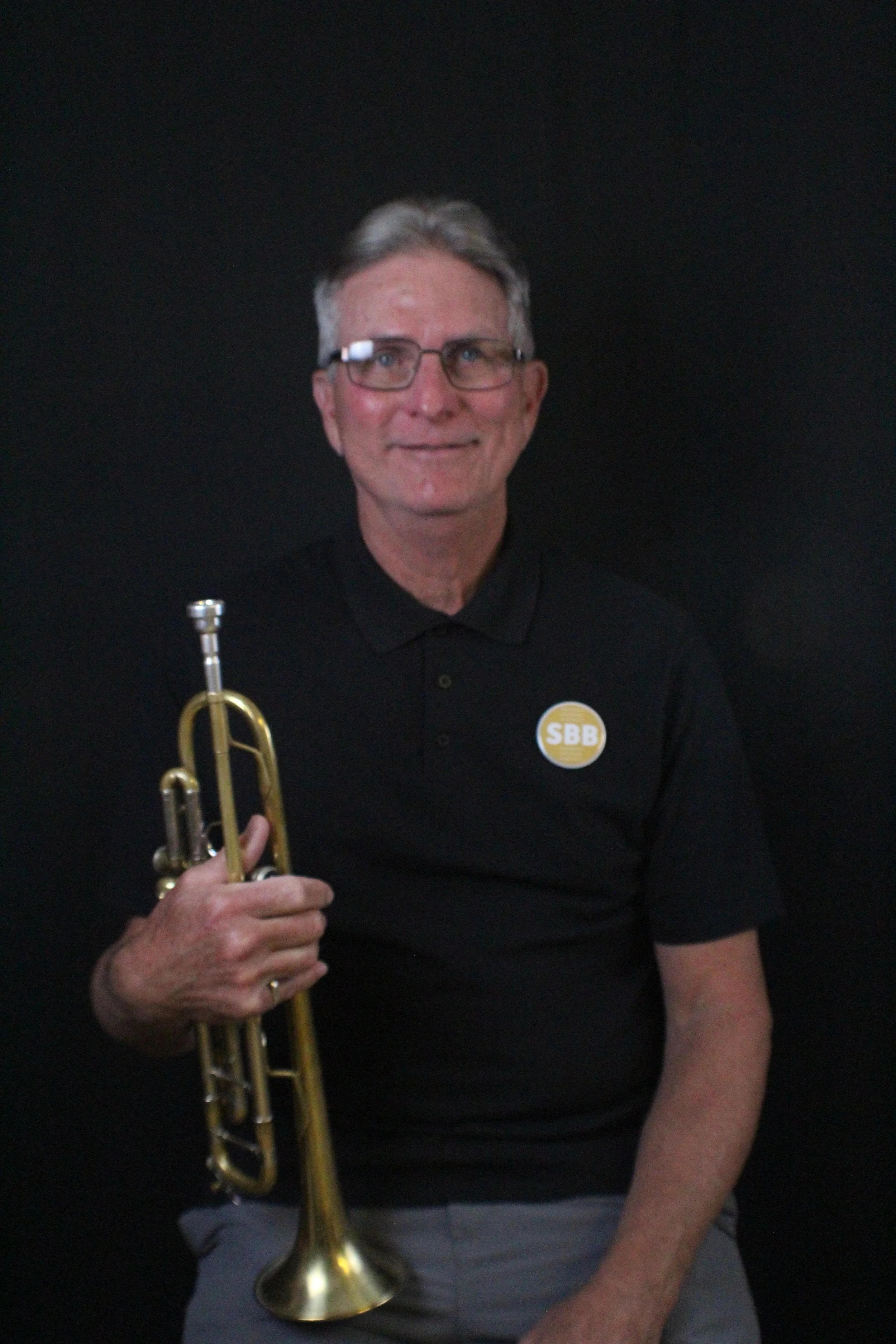 A middle-aged man with gray hair and a goatee smiling, holding a trumpet in his right hand, standing against a white brick wall.