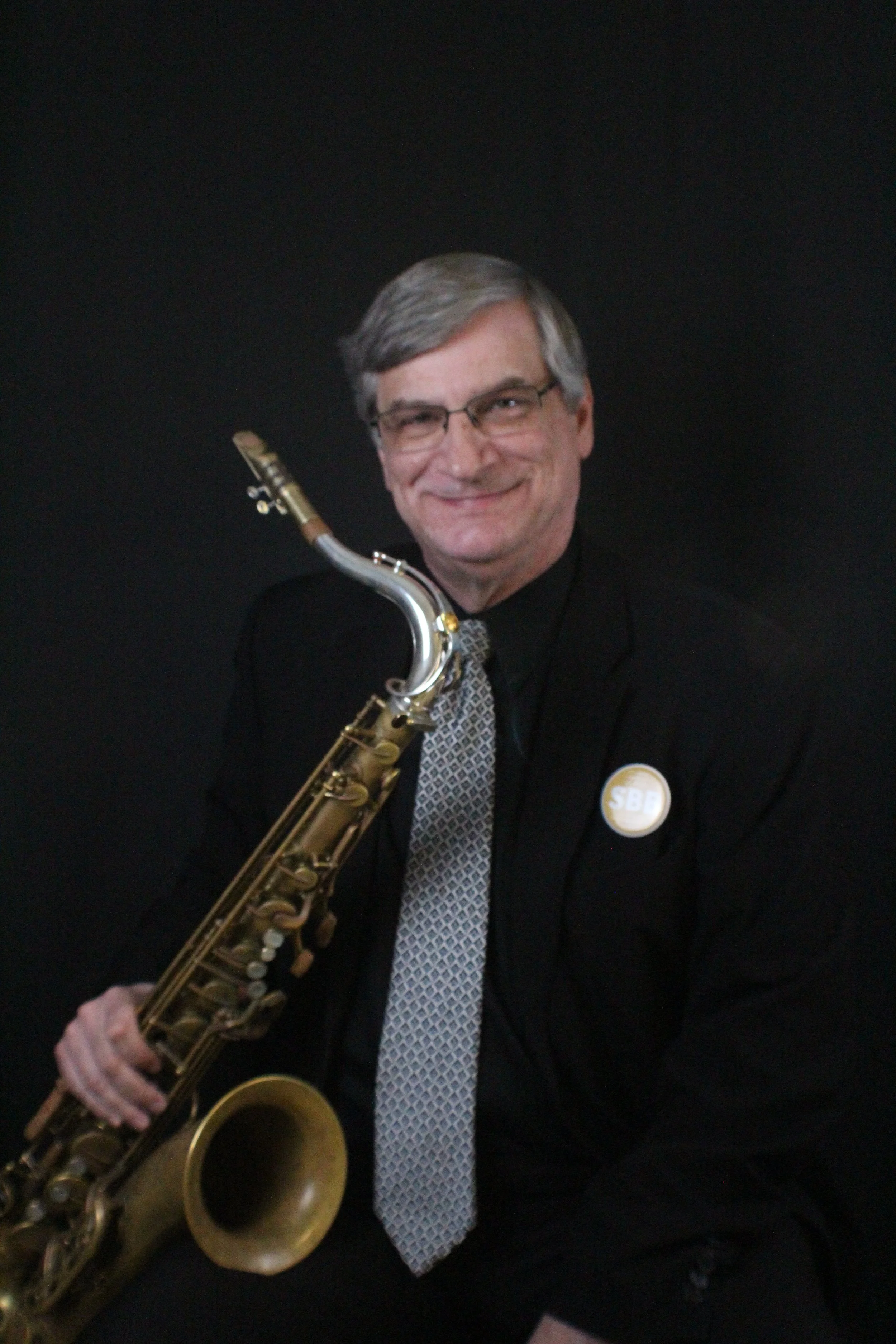 A middle-aged man with glasses and gray hair holding a saxophone, standing against a beige wall.
