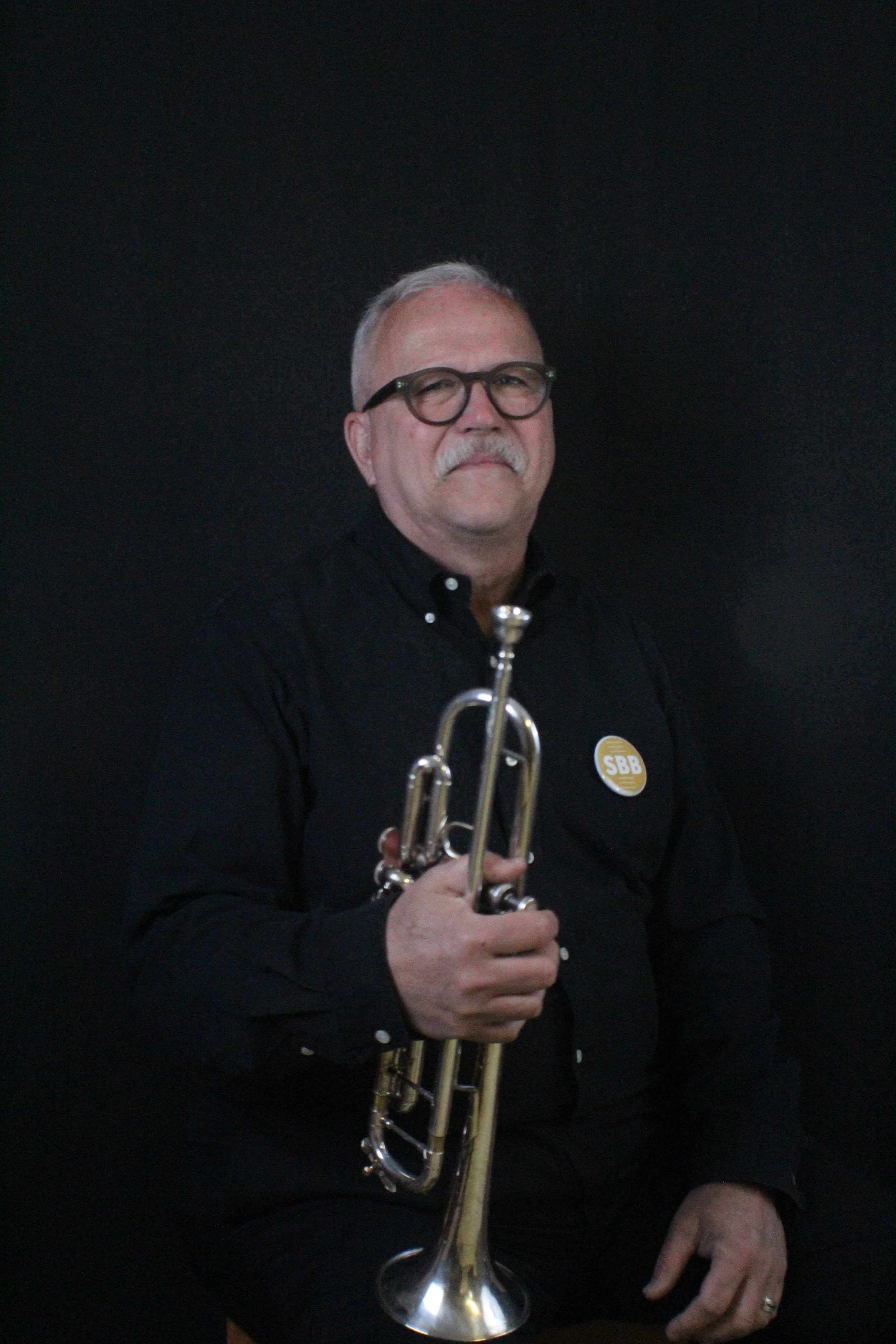 A smiling man with gray hair and glasses holding a trumpet, standing against a beige brick wall.
