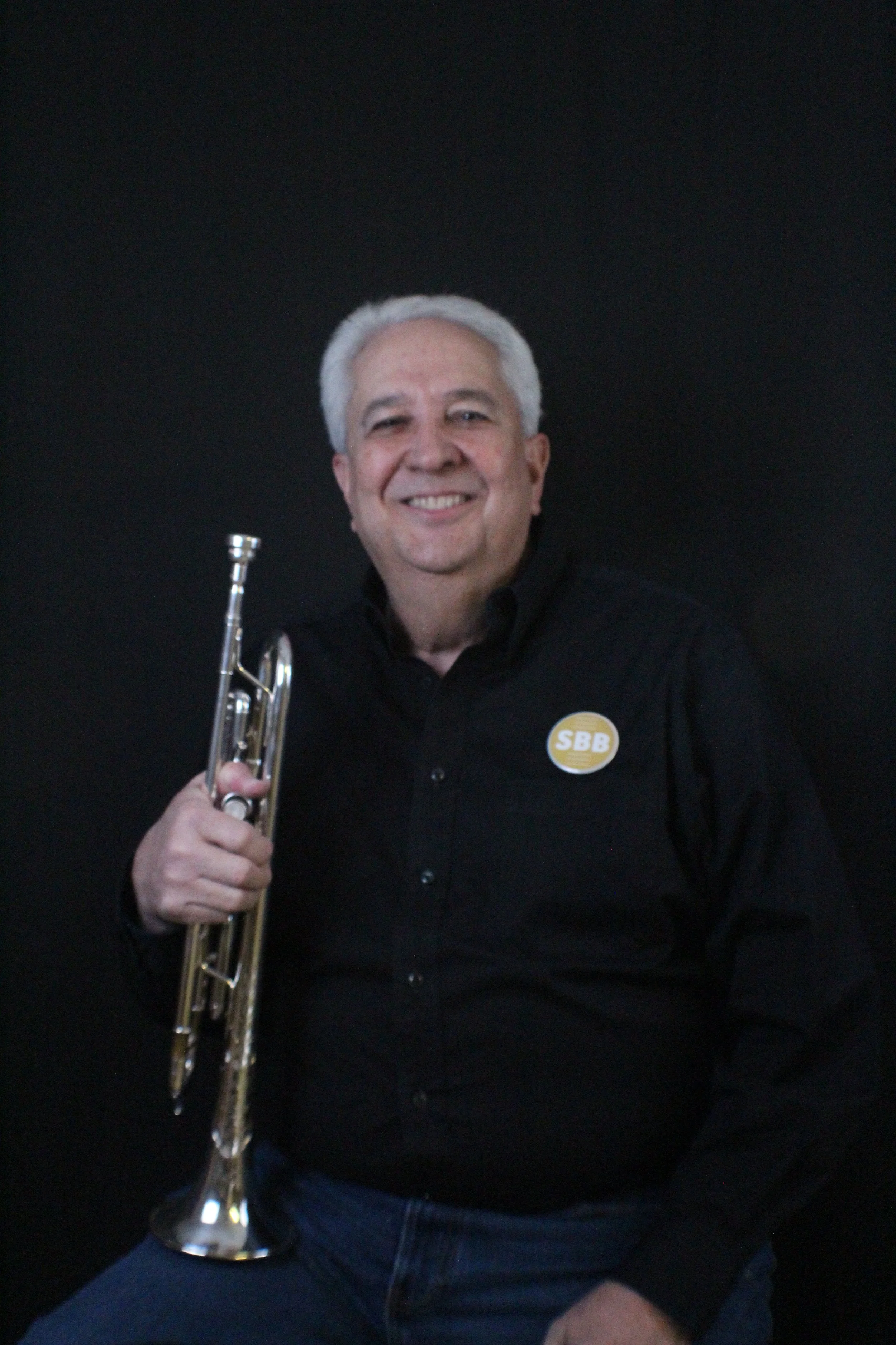 An older man with gray hair in a gray suit holding a trumpet, standing against a beige brick wall.