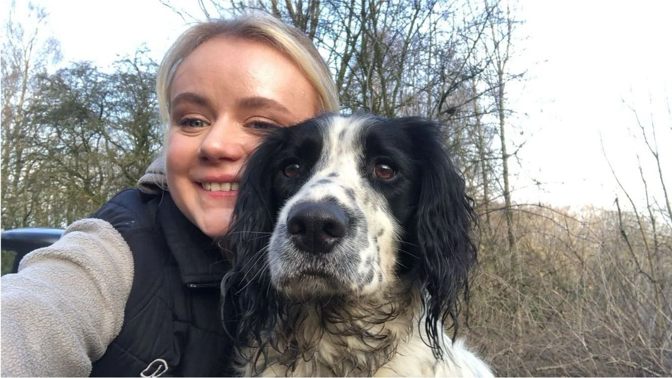 A woman taking a selfie with her black and white dog outdoors in a wooded area during daytime.