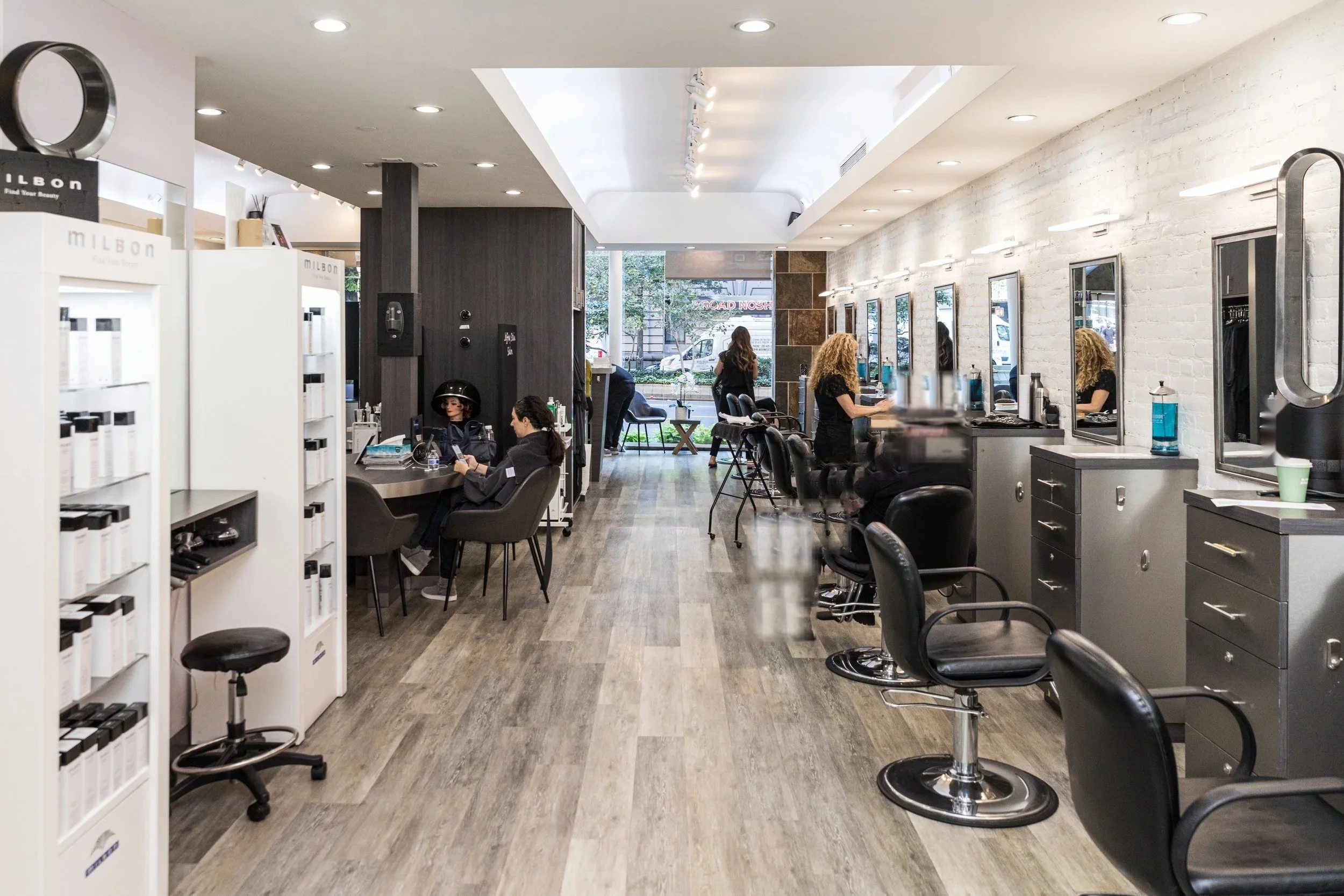 Interior of a modern hair salon with styling stations and clients getting hair services, large windows in the background letting in natural light.