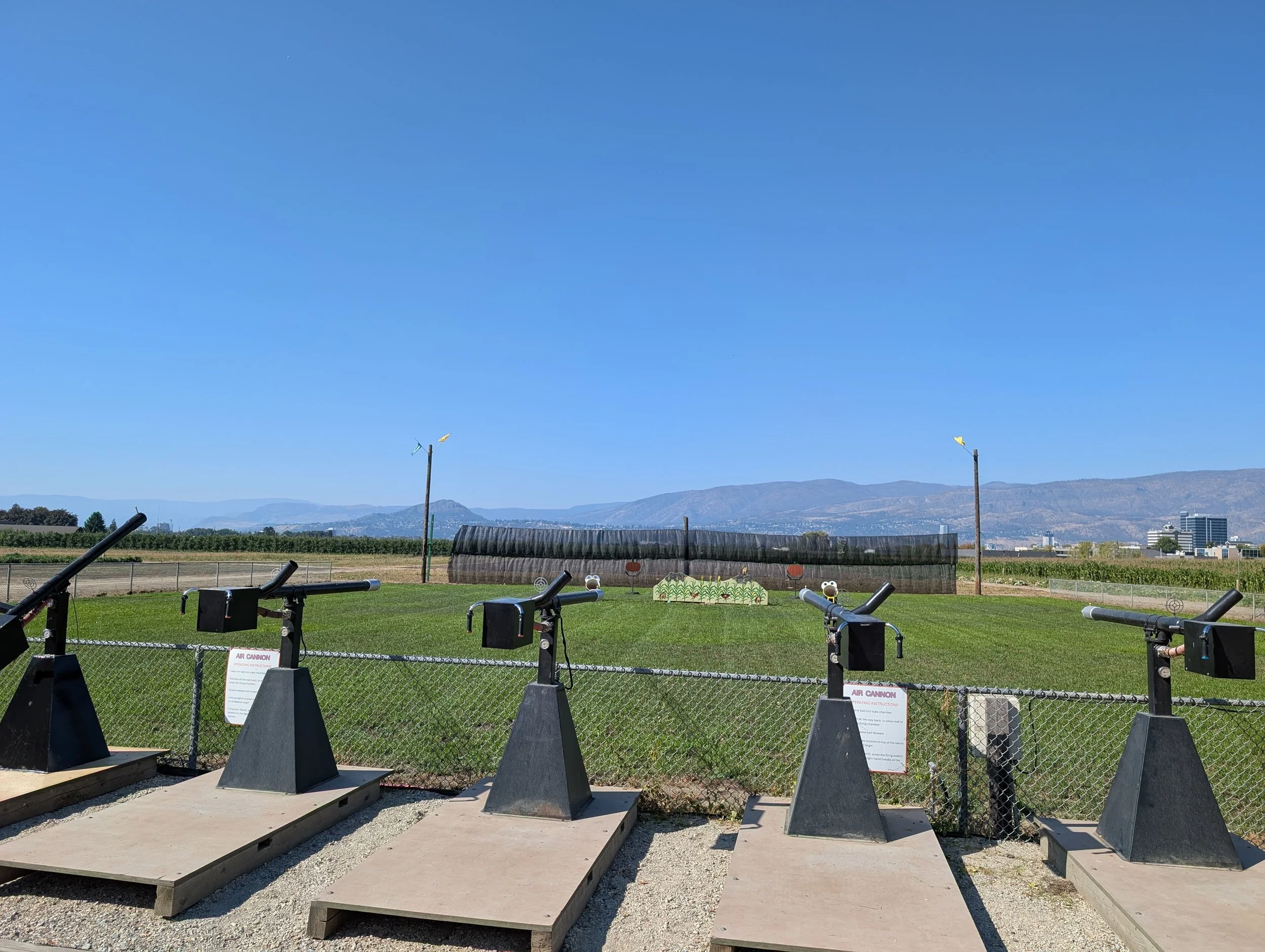 Open outdoor space on the farm with four air cannons aimed at a field with targets, fenced in, with a sign on the fence.