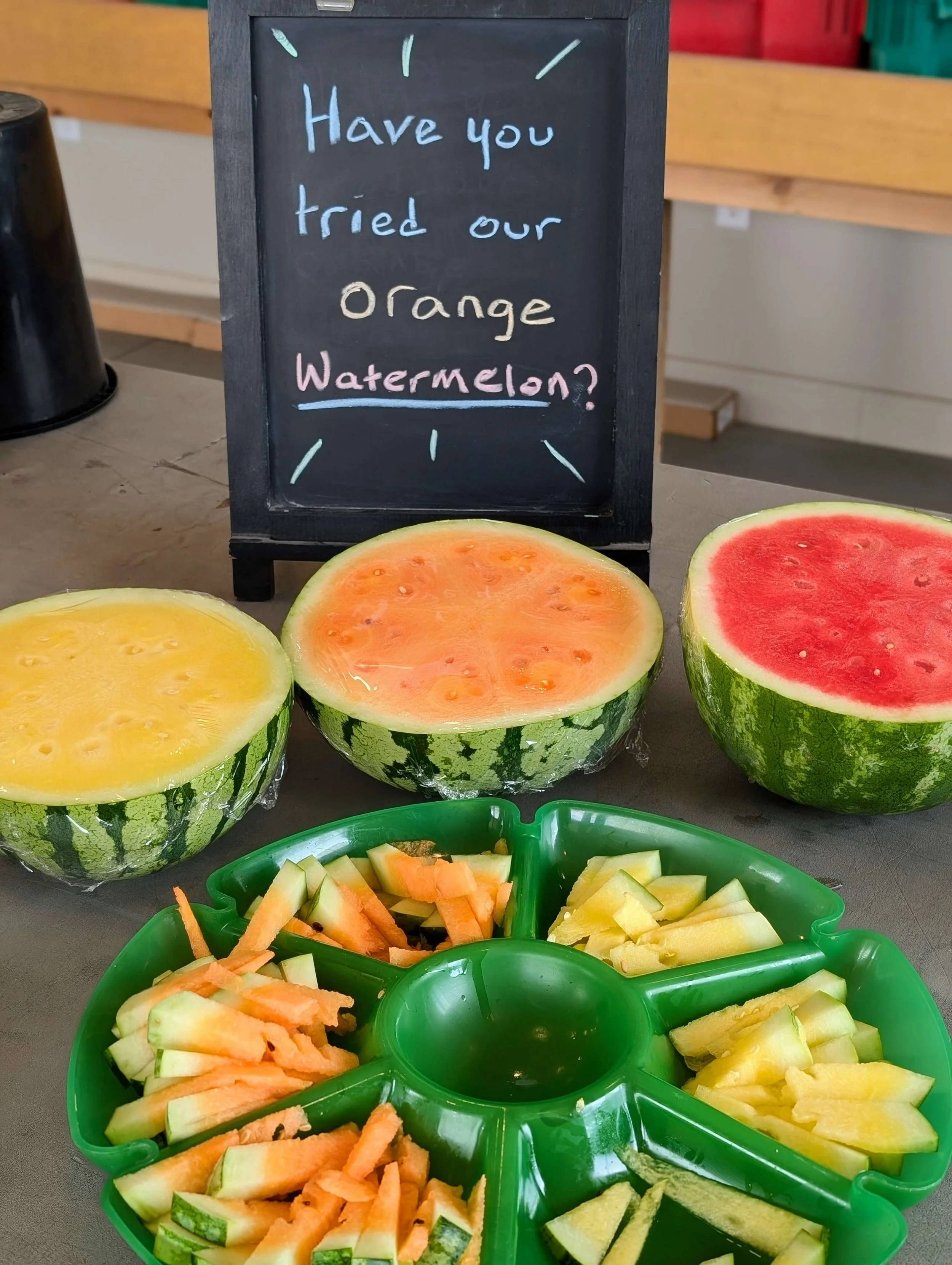 Watermelons cut in half to reveal yellow, orange, and red flesh, with a green plastic tray of sliced watermelon samples, and a blackboard sign that says, 'Have you tried our orange watermelon?'