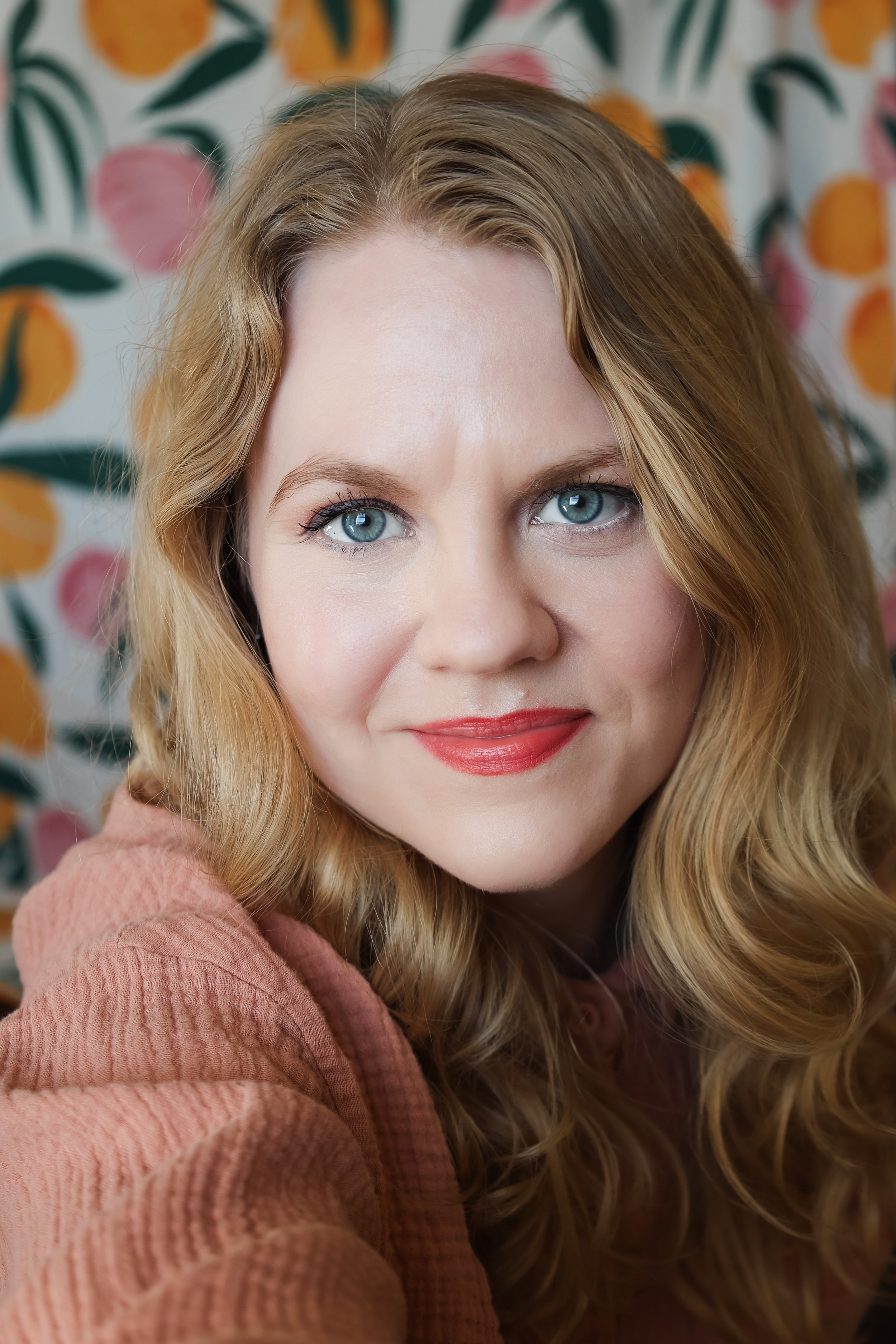 A woman with wavy blonde hair and blue eyes smiling at the camera, wearing a pinkish-brown top, with a colorful floral background.
