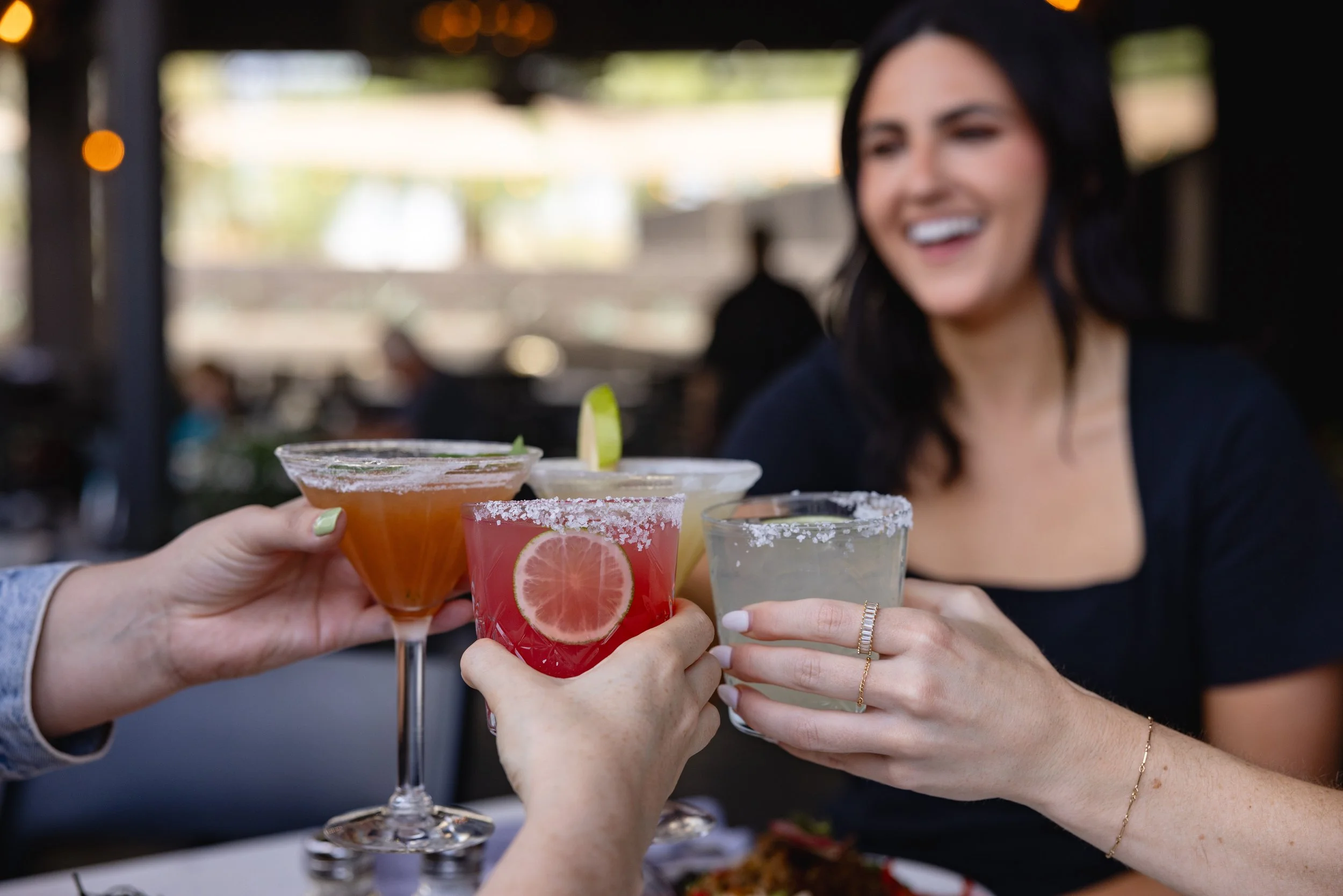 Friends toasting cocktails including a Blood Orange Margarita, Thirsty Lion Margarita, and martinis, capturing a lively happy hour atmosphere with handcrafted drinks and social energy.