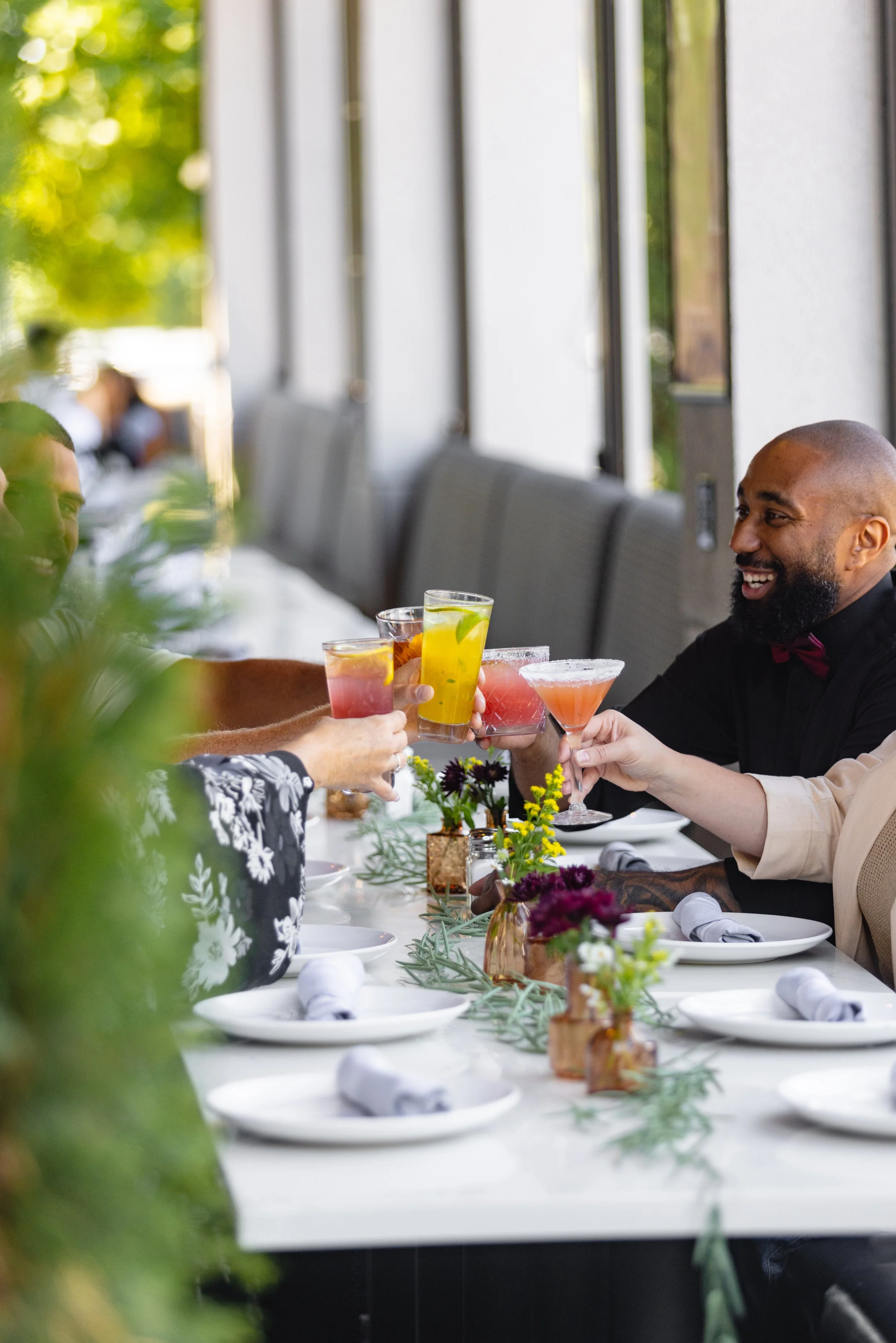 Group of friends toasting colorful cocktails at a bright, modern dining table, showcasing a lively atmosphere ideal for group dining events, happy hours, and celebrations.