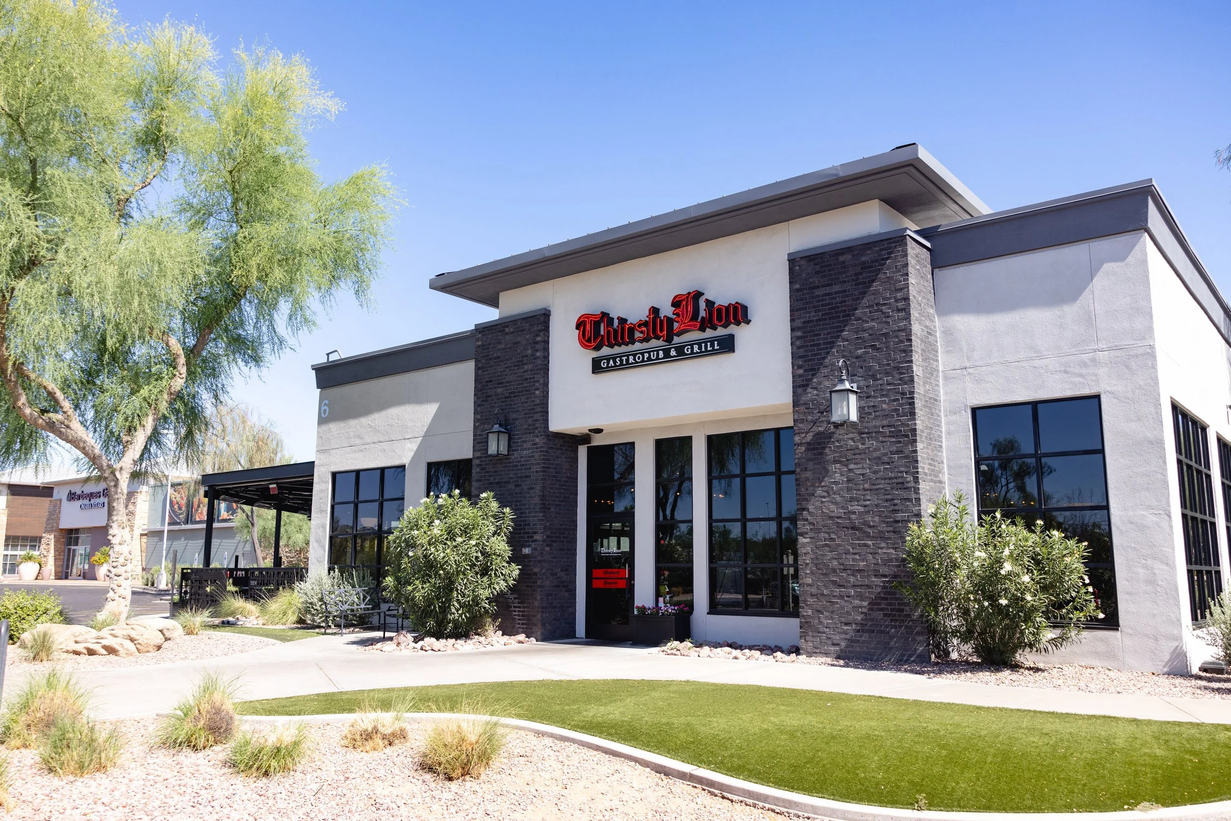 Exterior of Thirsty Lion Desert Ridge location restaurant with black walls, large windows, and an inviting outdoor seating area with trees and a small lawn.