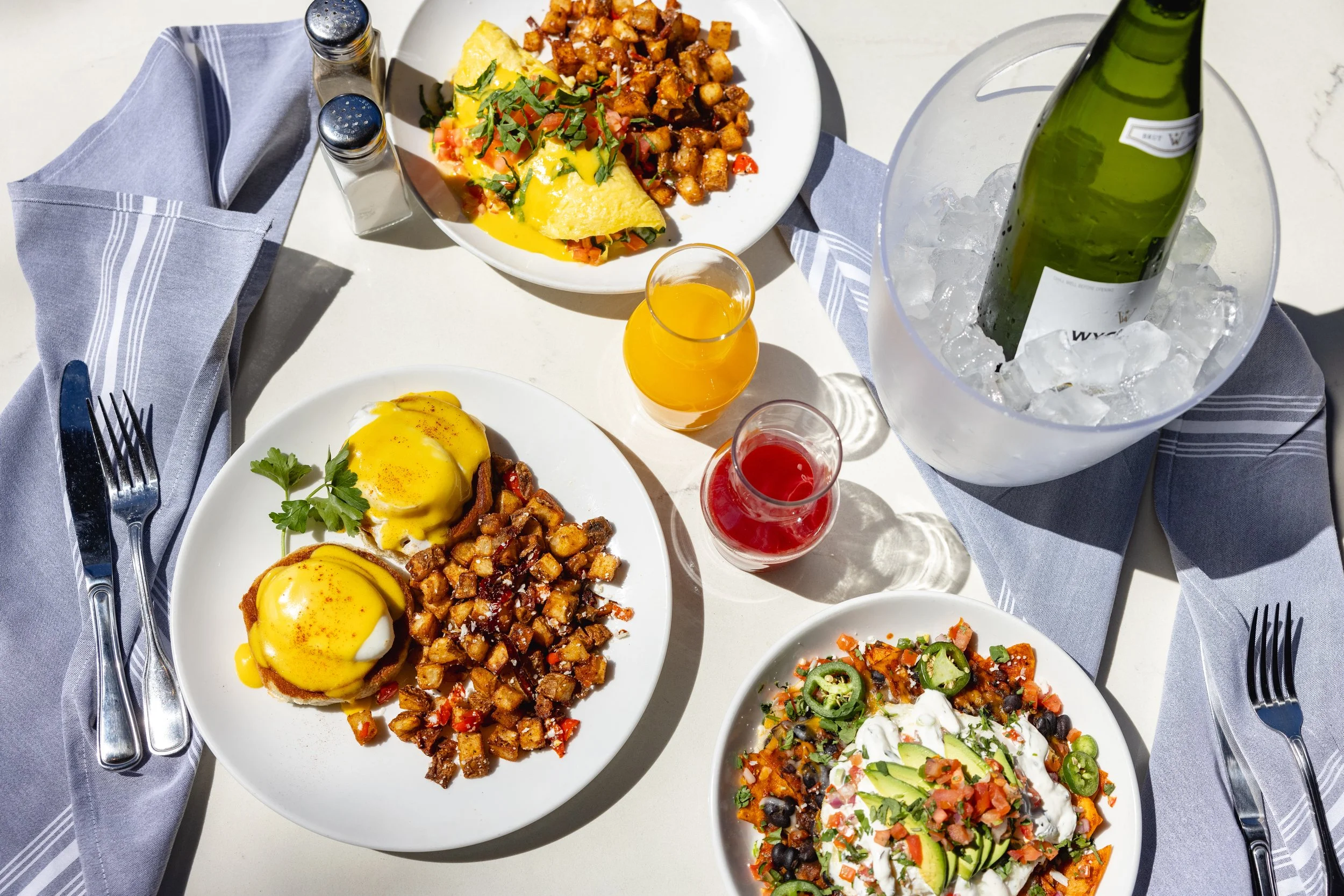 Overhead view of weekend brunch spread featuring Eggs Benedict, spinach and feta omelette, Spanish chilaquiles, and Mimosa by the Bottle with fresh juices, highlighting a vibrant brunch experience with food and cocktails.