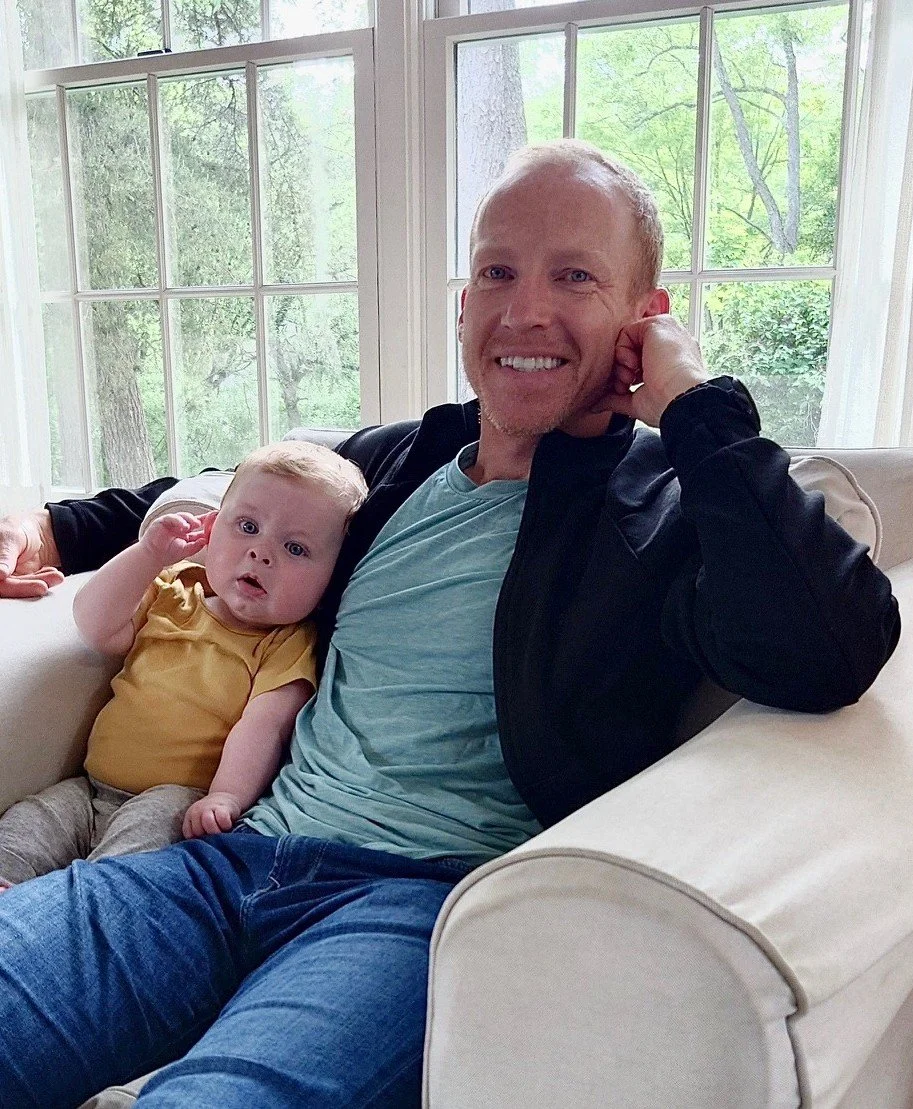 A man sits smiling with a baby on his lap in an armchair.