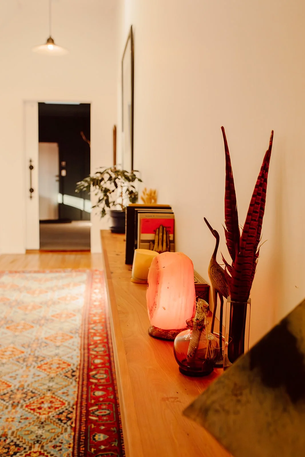 A wooden console table decorated with a Himalayan salt lamp, a small decorative bowl, a salt shaker, and a vase with red and purple leaves. Behind the table are some books and a potted plant. The space is warmly lit with a hanging ceiling light in th