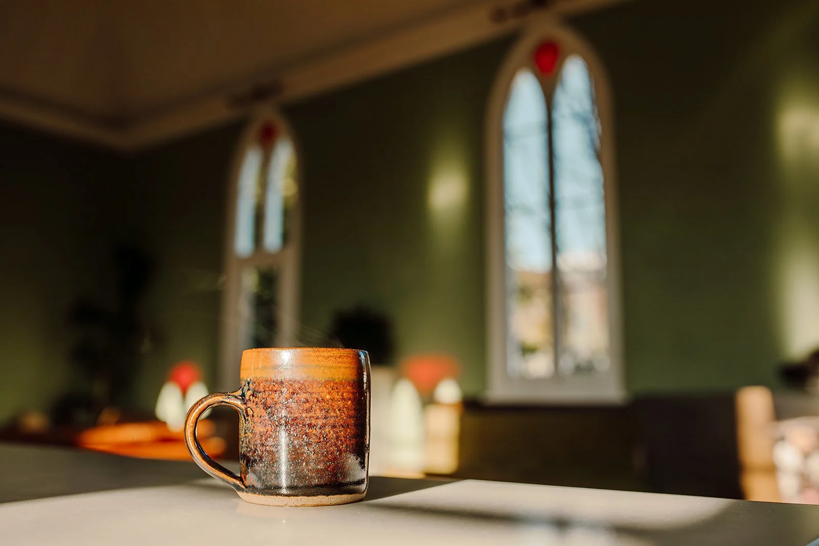 A glazed ceramic mug on a table in a sunlit room with green walls and tall arched windows.
