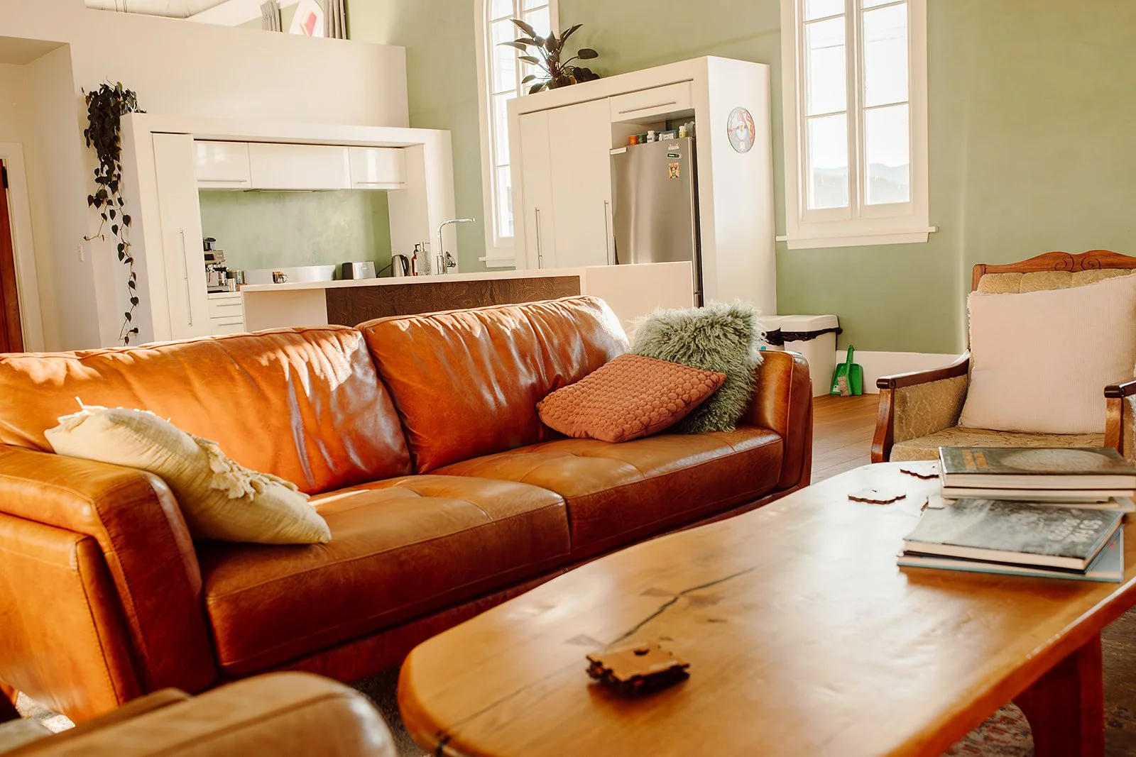 Living room with a leather sofa, pillows, a wooden coffee table with books, and an armchair. The background features a kitchen with white cabinetry, a refrigerator, and windows with daylight streaming in.
