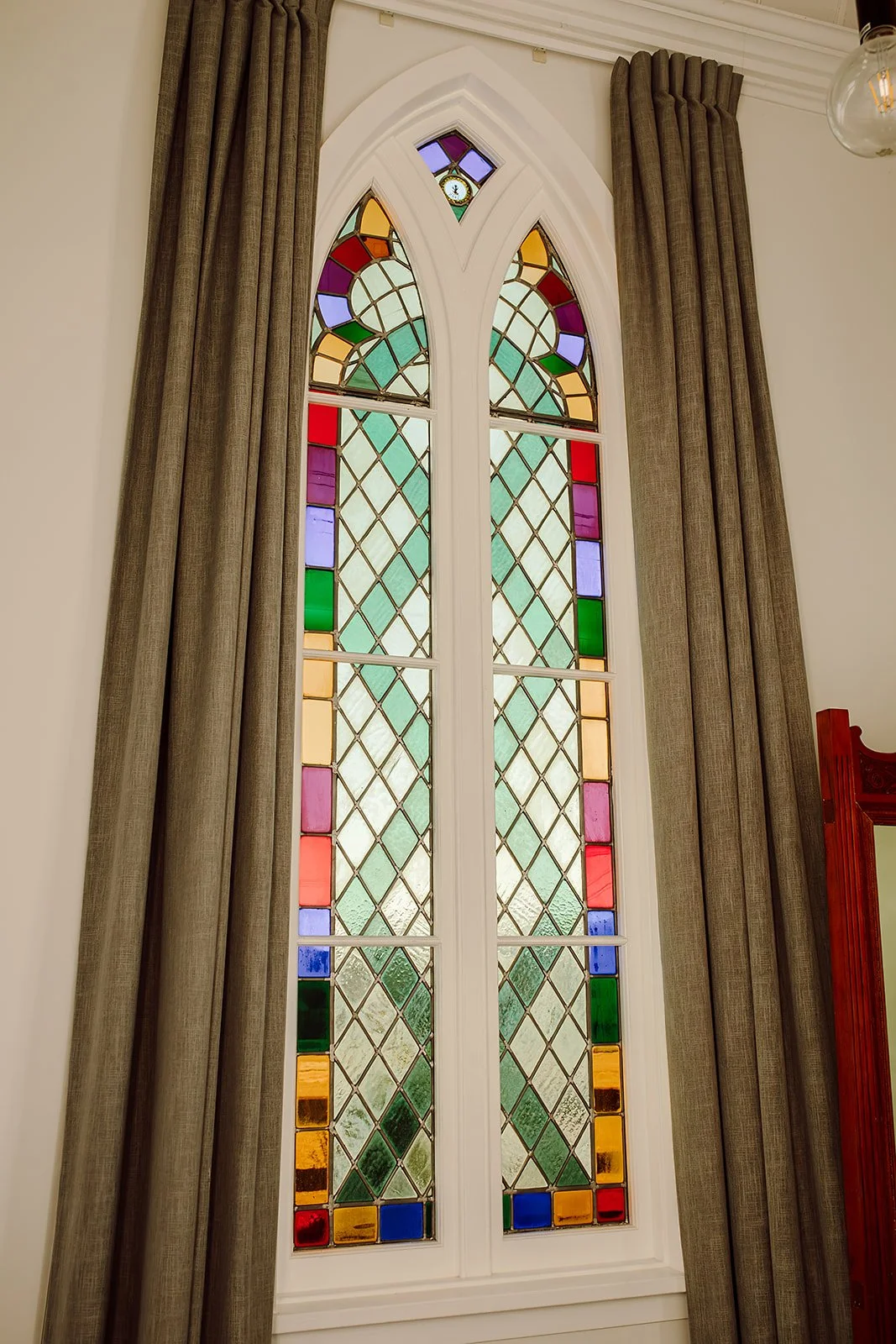 Stained glass window with geometric patterns and colorful glass panes, framed by beige curtains in a room.