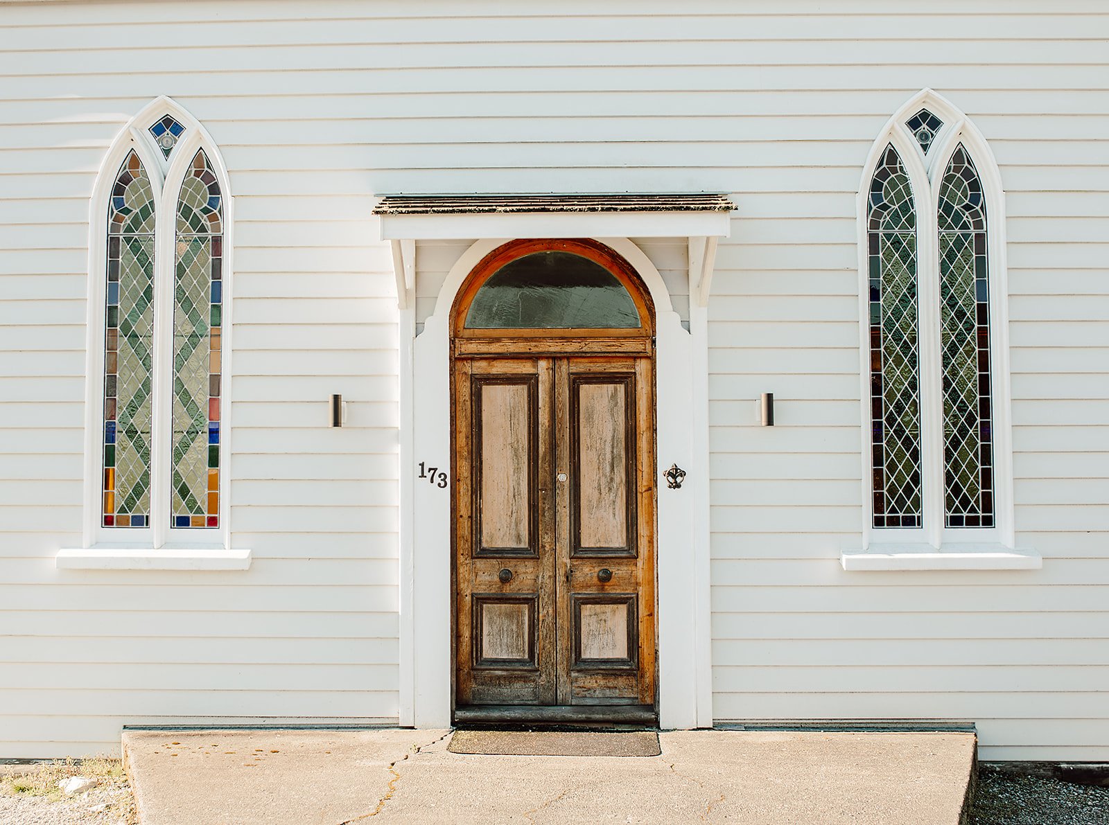 Front view of a white church with a wooden double door and two tall stained glass windows on each side.