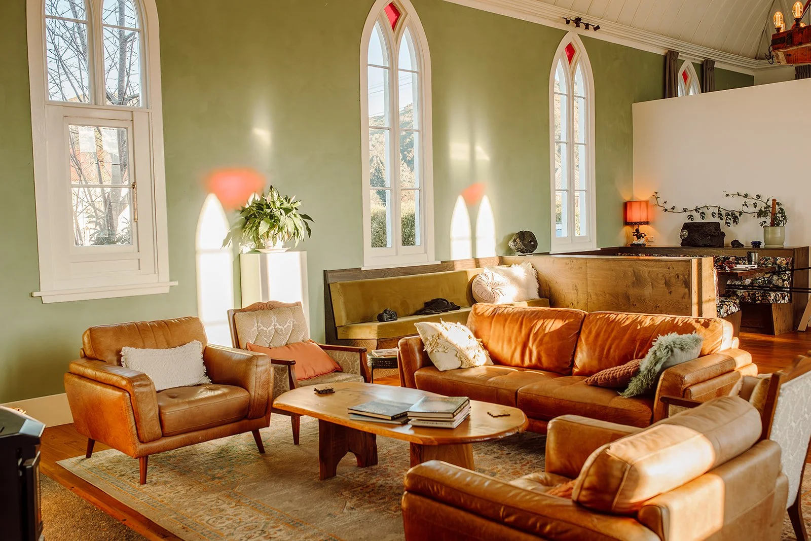 A cozy living room with green walls, large arched windows, and a variety of seating including leather armchairs and a sofa, with a wooden coffee table, lamps, and decorative cushions.