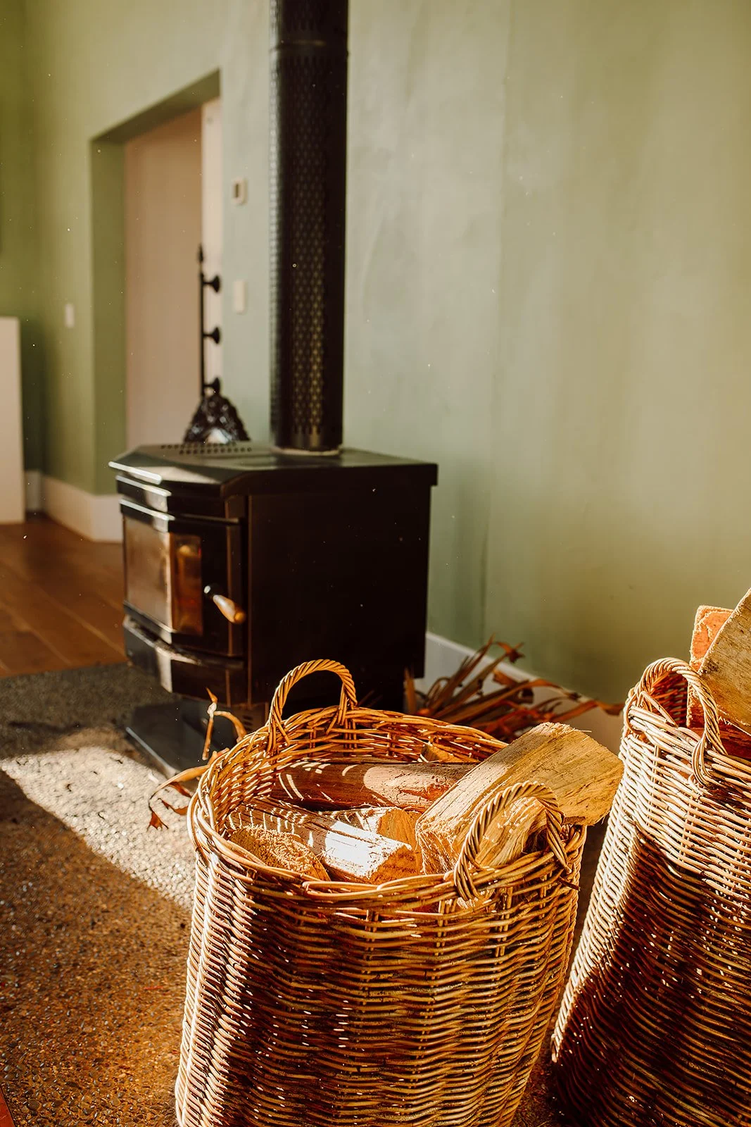 A cozy indoor scene with a black wood-burning stove, wicker baskets filled with chopped firewood, and a green wall in the background.