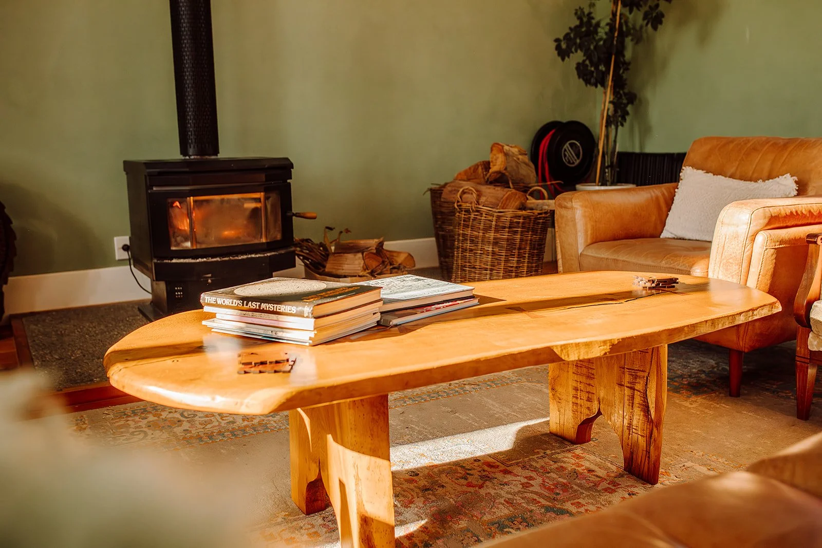 Living room with a wood-burning stove, a wooden coffee table with books on it, a leather sofa with a white pillow, a wicker basket filled with firewood, and a green wall.