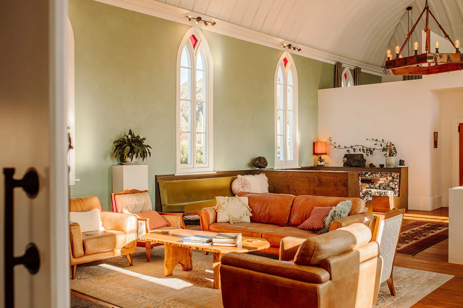 Living room with three tall arched windows, a green accent wall, and vintage leather furniture including a sofa and armchairs, wooden coffee table, and decorative pillows. There is a chandelier hanging from the ceiling and a small table with a lamp o