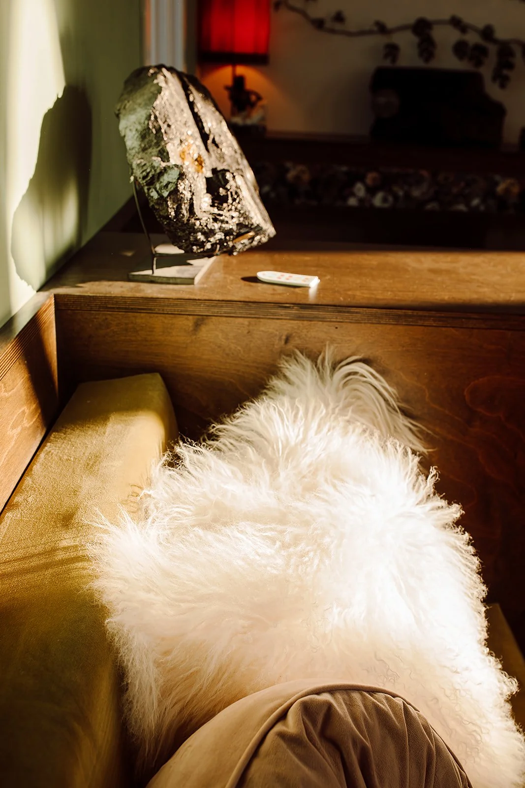A cozy living room corner with a cream-colored furry blanket or cushion on a wooden chair or couch. In the background, there's a wooden table with a decorative black and gold mask on a stand, a remote control, and a lamp with a red shade. The room ha