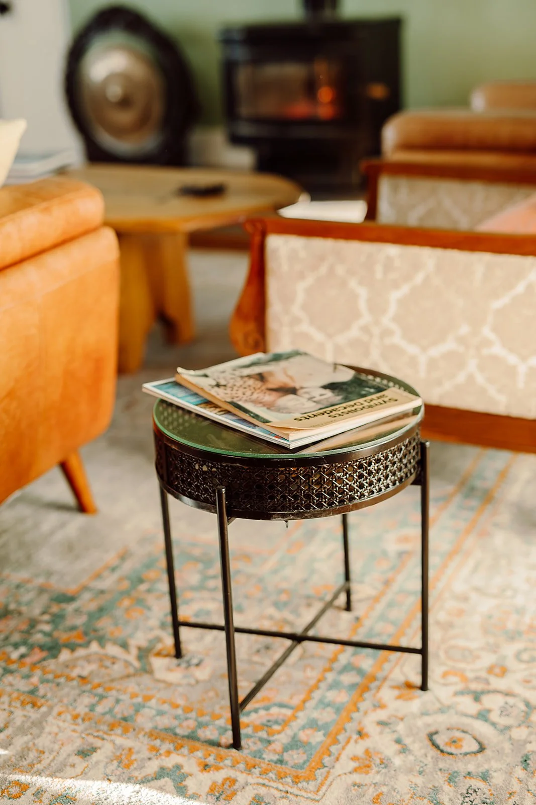 A small round black metal table with a glass top, placed on a patterned rug, holding magazines in a cozy living room.